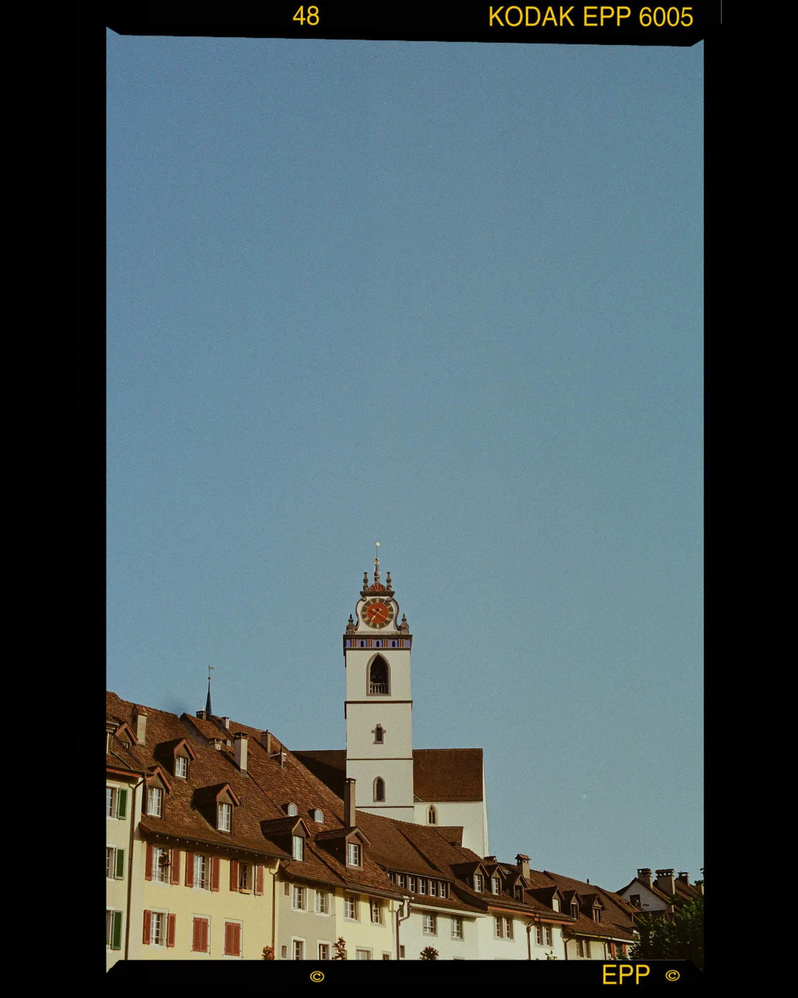 A church tower with a clock rising above old rooftops against a clear blue sky, the scan showing its film border.
