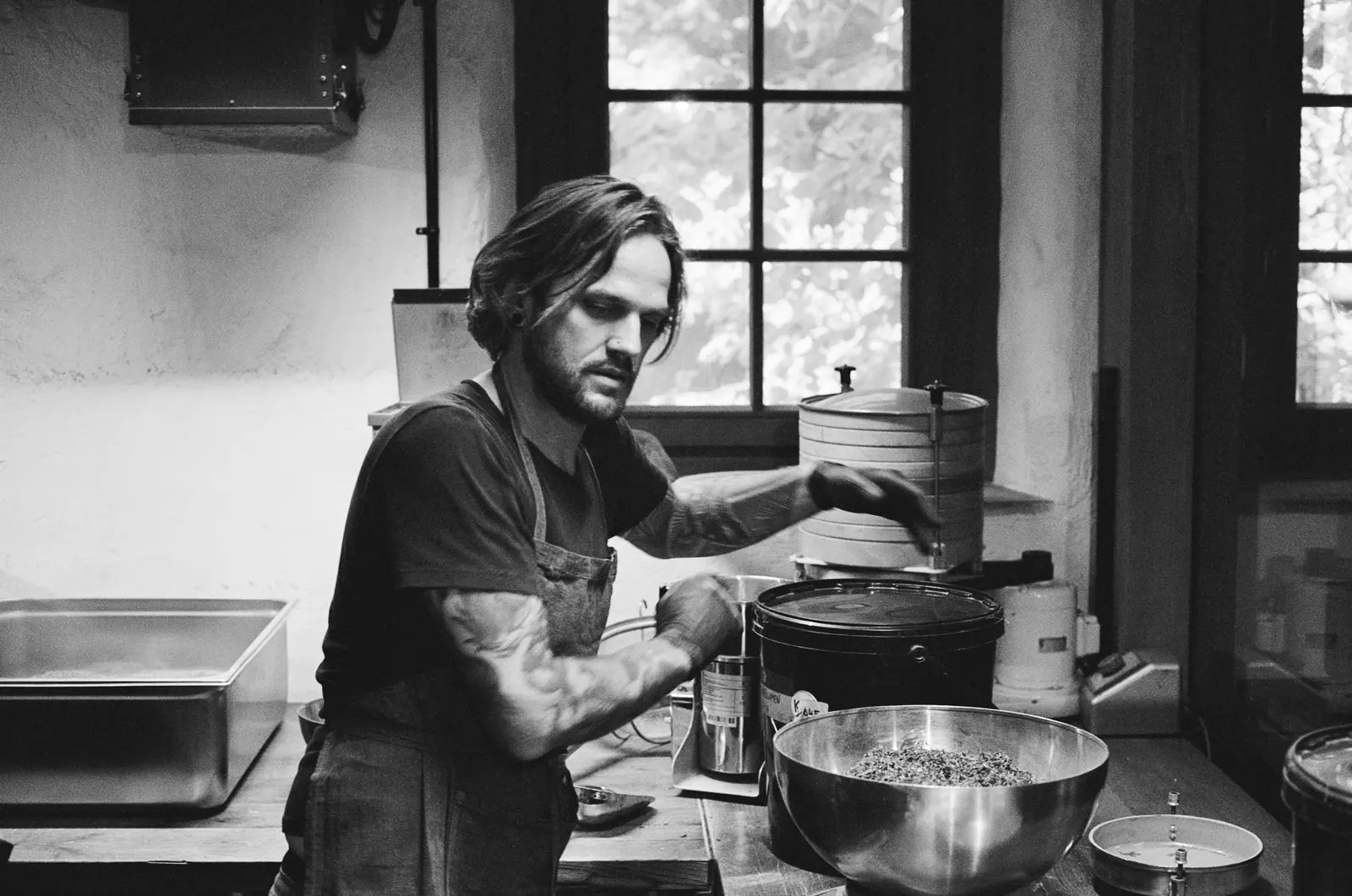 Black-and-white: a tattooed man in an apron cooks at a steel counter by a window.