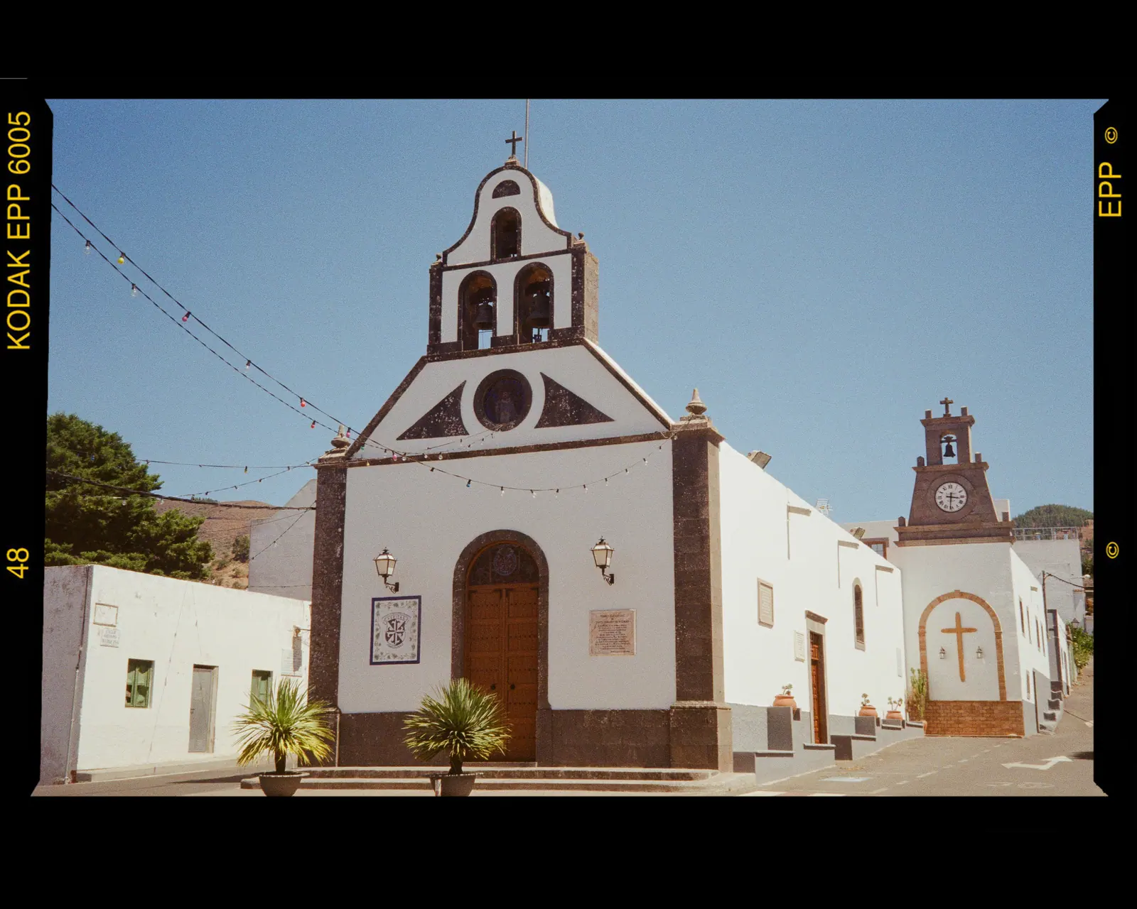 A white chapel with a bell gable and a smaller clock tower beside it under a blue sky, framed by the film border.