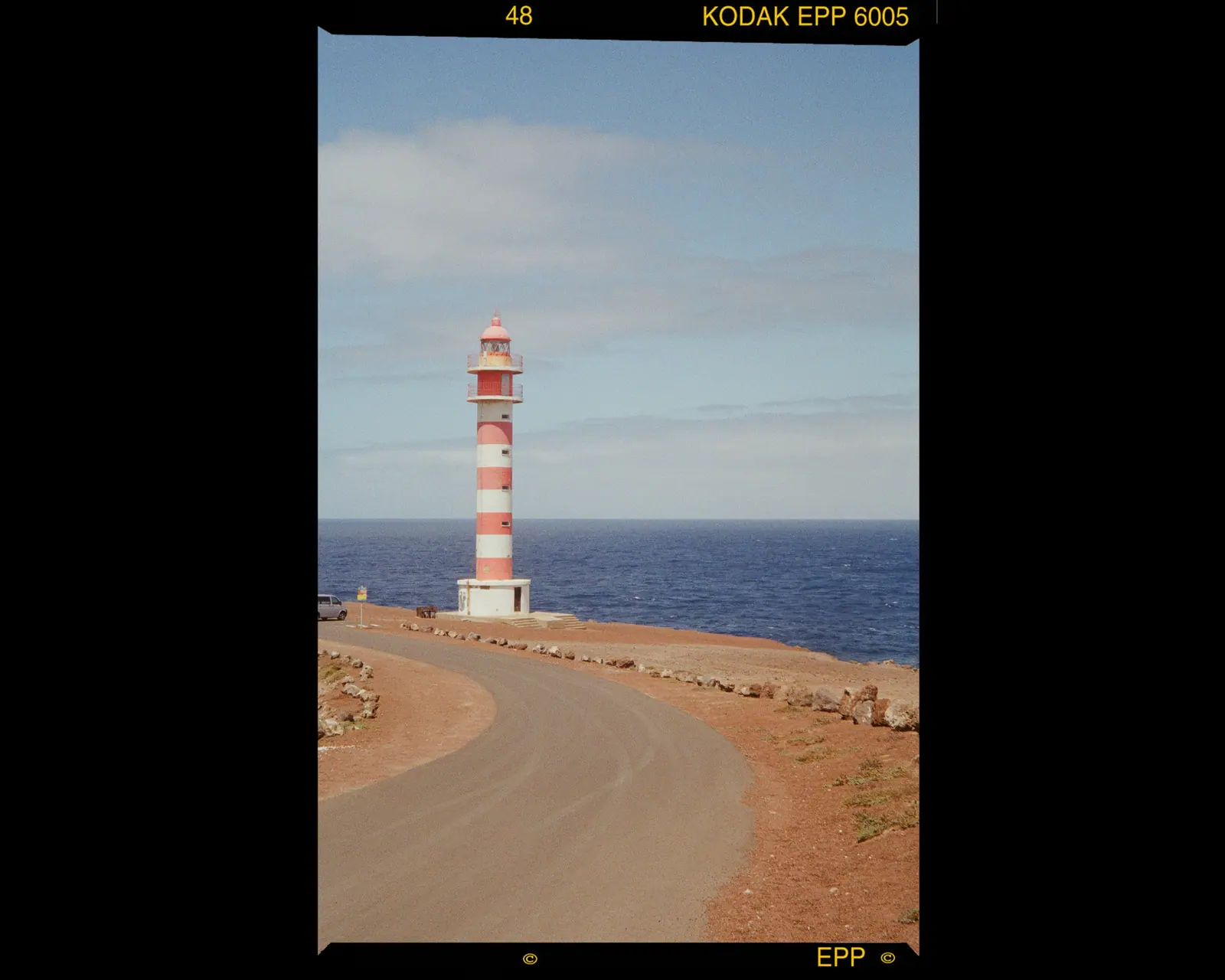 A red-and-white lighthouse at the end of a dirt road by a deep blue sea, framed by the film border.