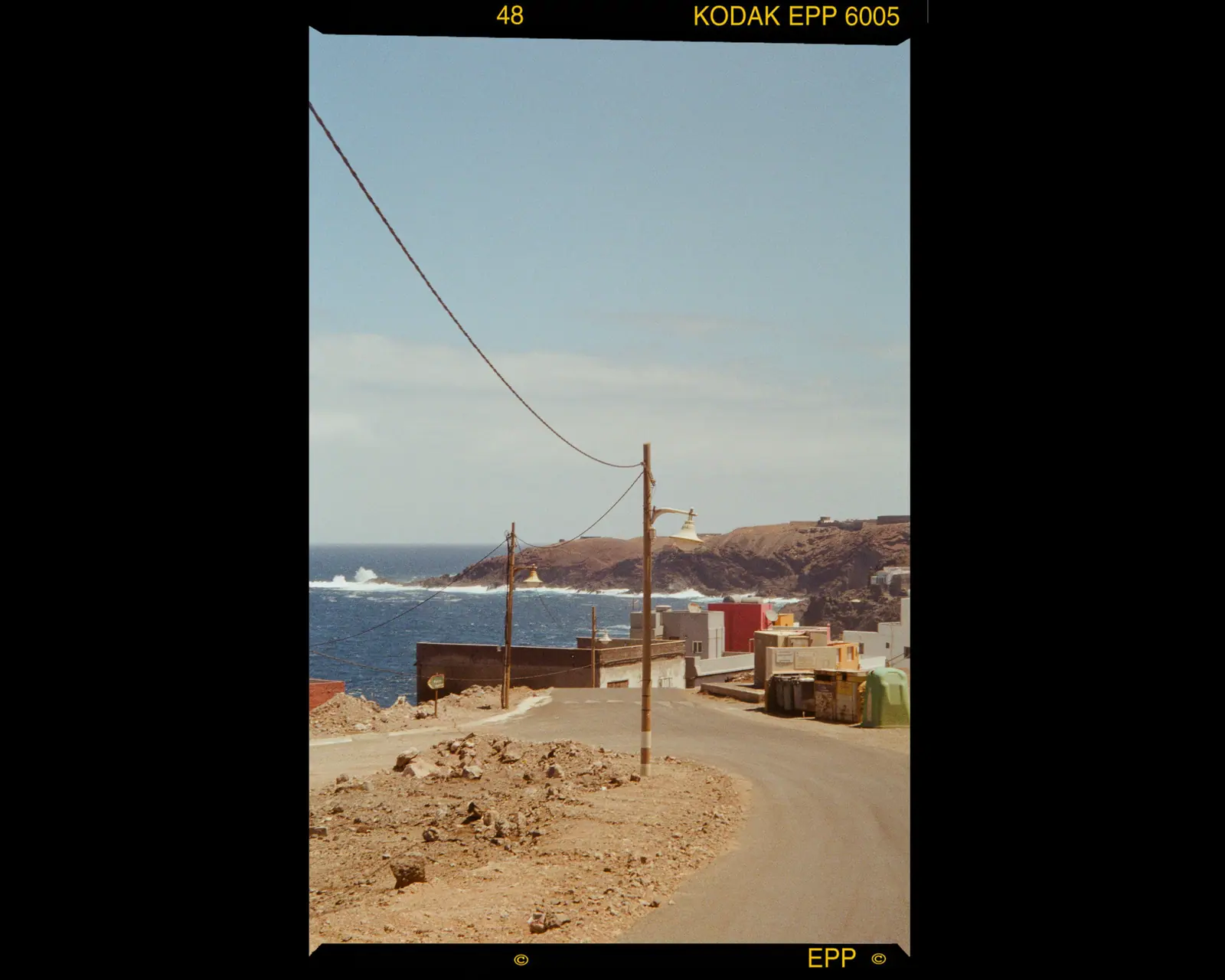 A dusty coastal road with a leaning power line running toward low houses and the sea, framed by the film border.