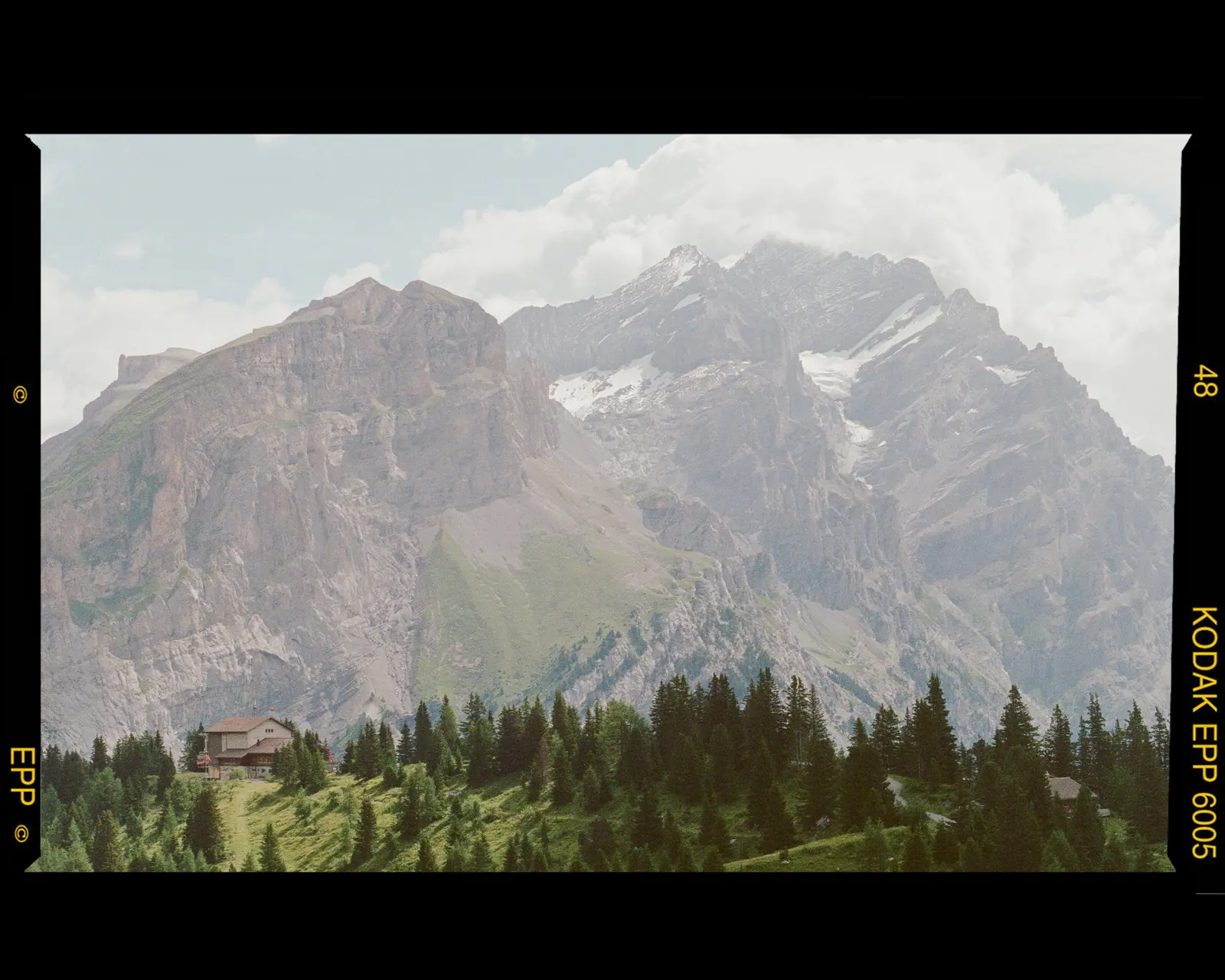 A rugged limestone massif above a band of dark forest and a small chalet, framed by the film border.