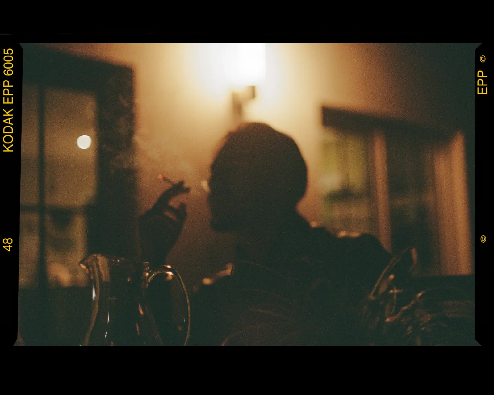 A figure in silhouette smoking in warm lamplight, glasses on the table in the foreground.