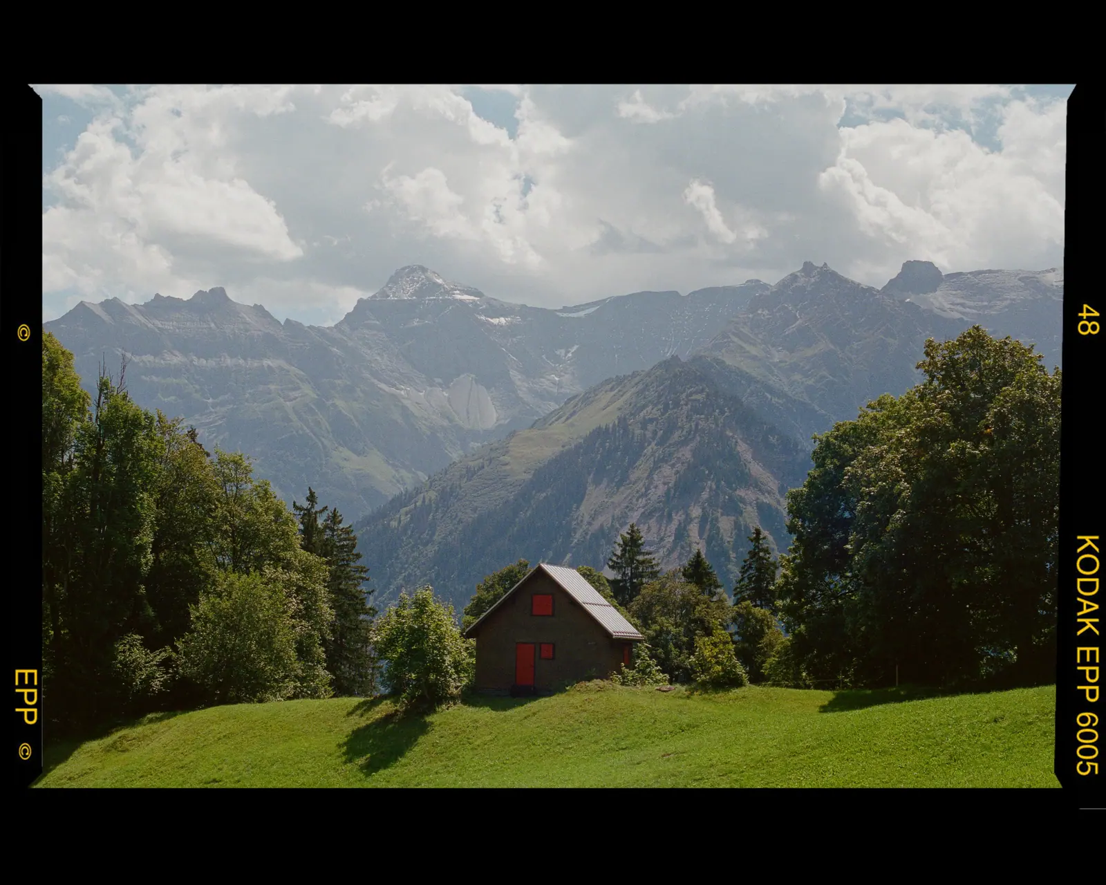 An alpine valley with a small red-roofed hut in a meadow, trees and a mountain range under drifting clouds.