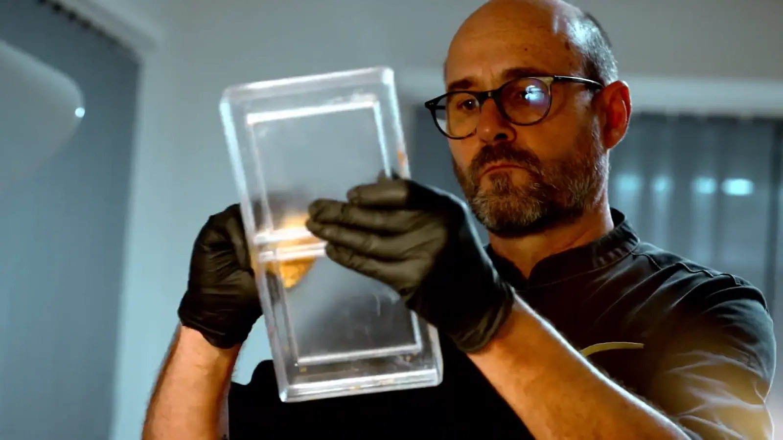 Still from the documentary: a man in glasses and black gloves holds a clear chocolate mould up to the light and examines it.