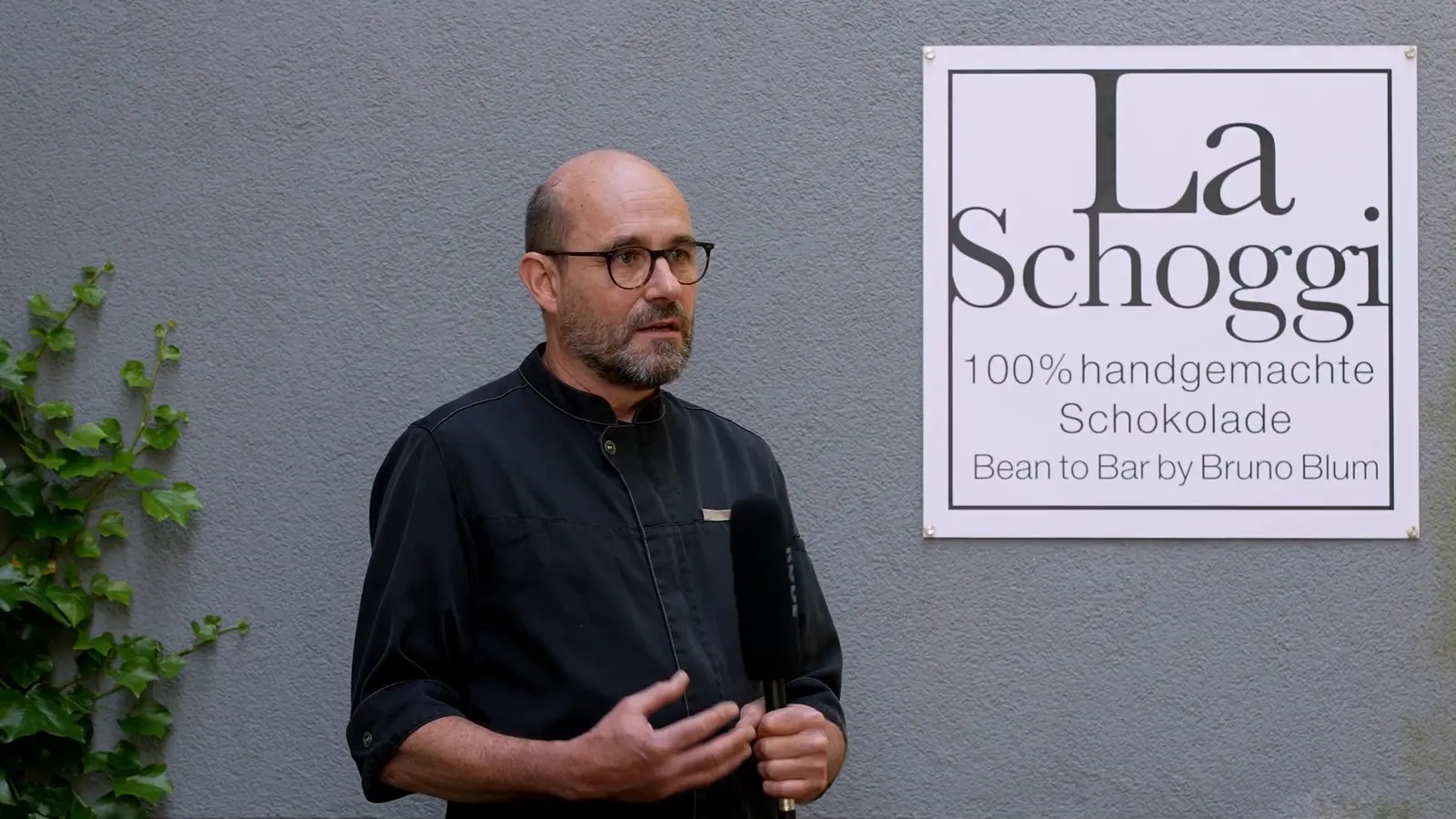 Still from the documentary: a man in glasses and a dark shirt speaks into a microphone in front of a shop sign reading “La Schoggi”.