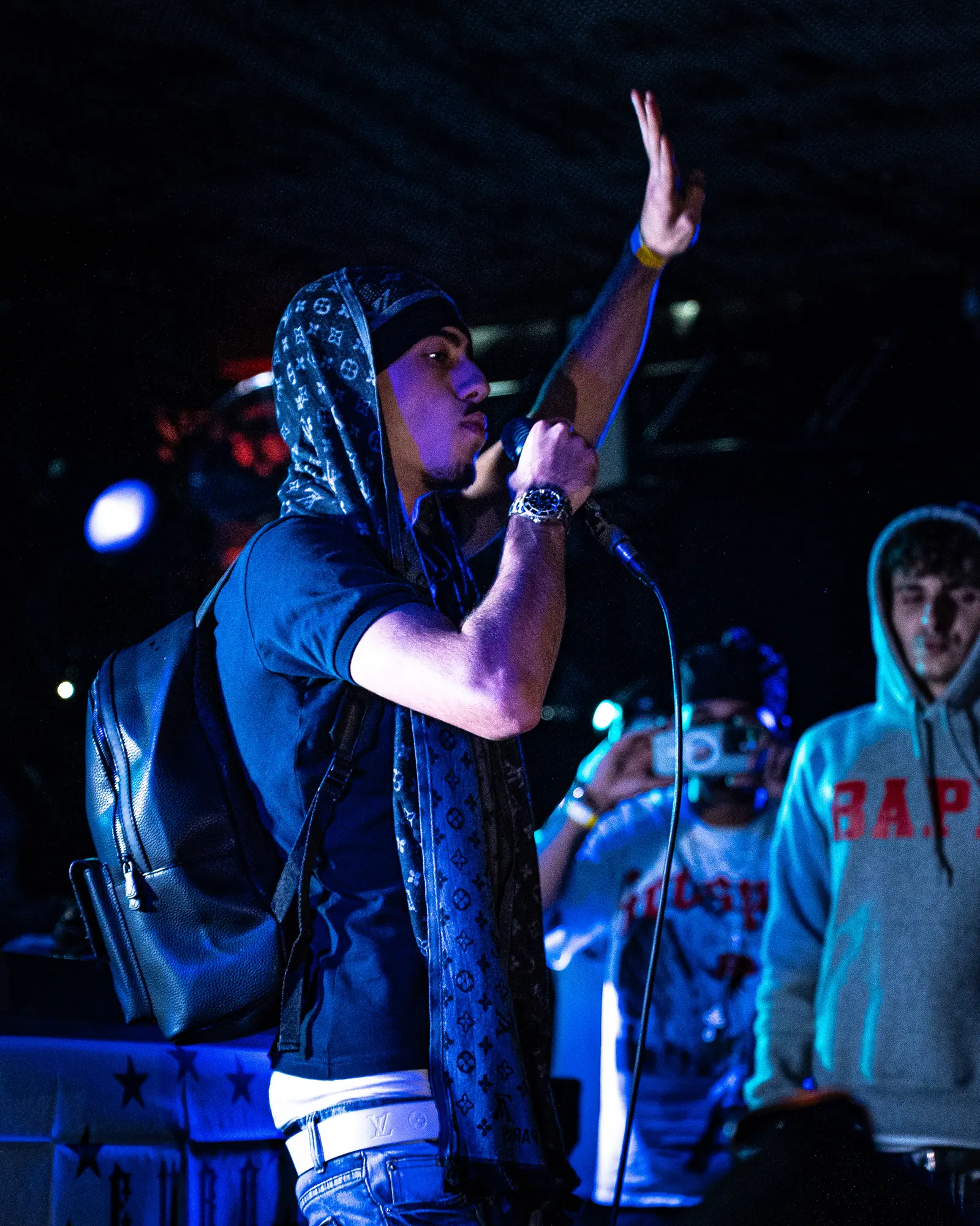 A rapper in a hooded jacket and patterned scarf raises one hand while performing, fans in hoodies behind him under blue stage light.
