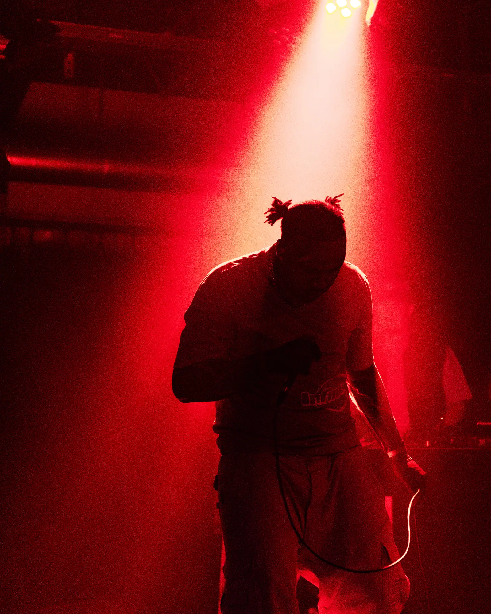 A performer in silhouette against a deep red stage light, head bowed over the microphone.