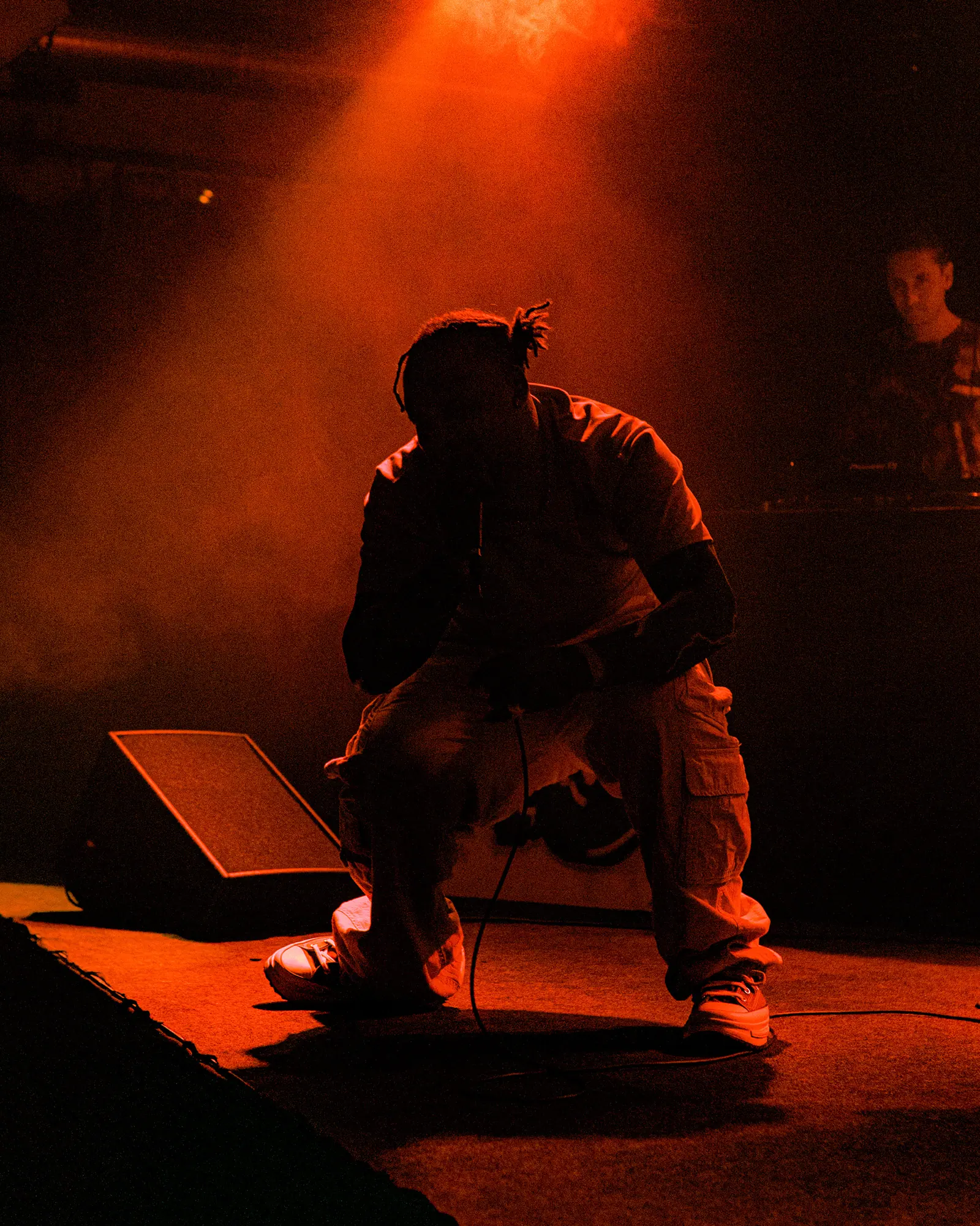 A performer crouches at the edge of the stage in orange-red light and smoke.