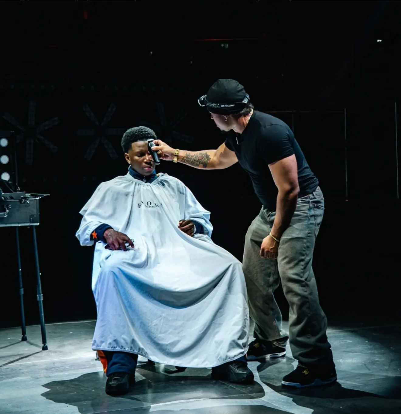 On a dark stage, a barber works on a man seated under a white cape.