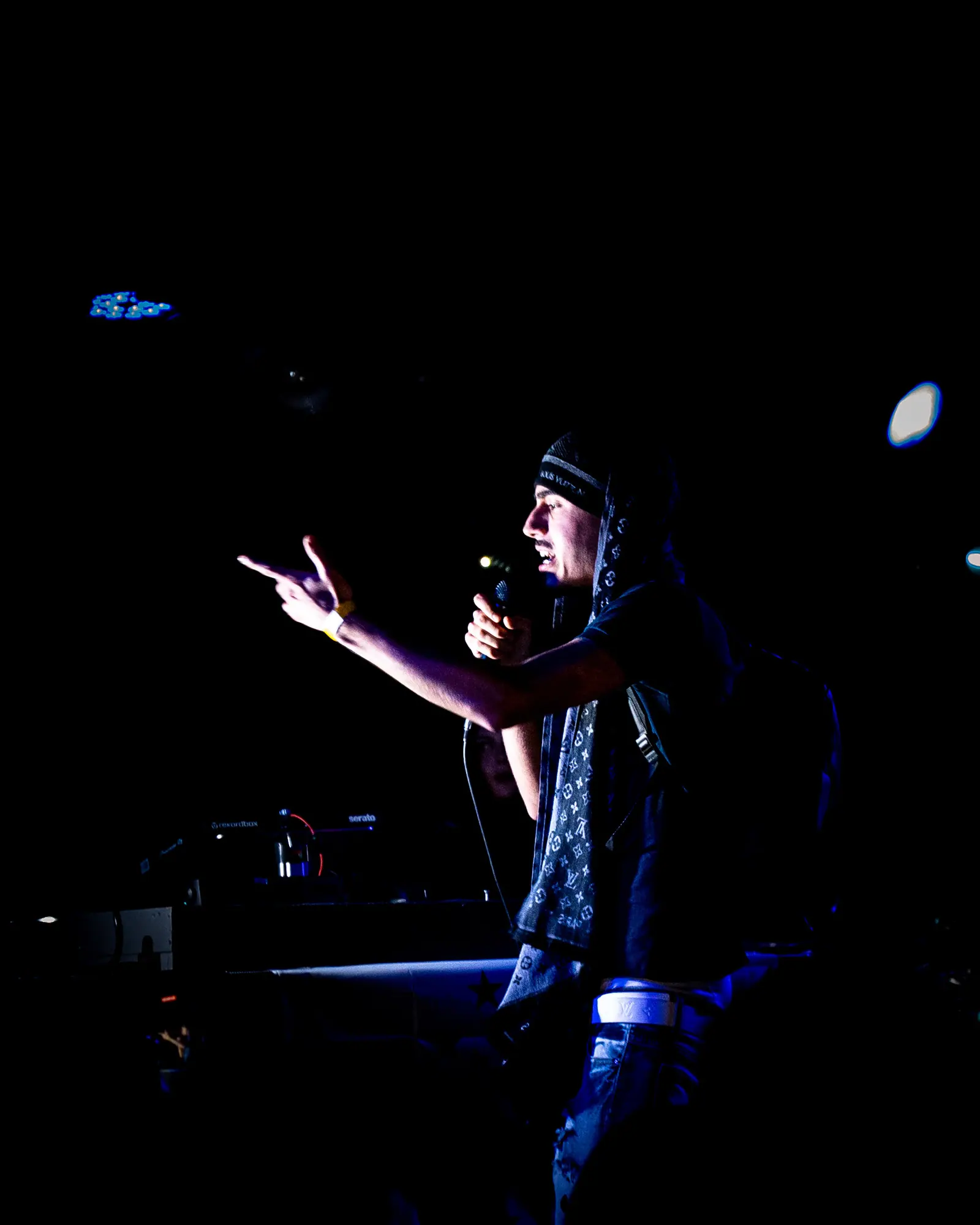A performer in a bandana points toward the crowd from a dark stage, microphone in hand.
