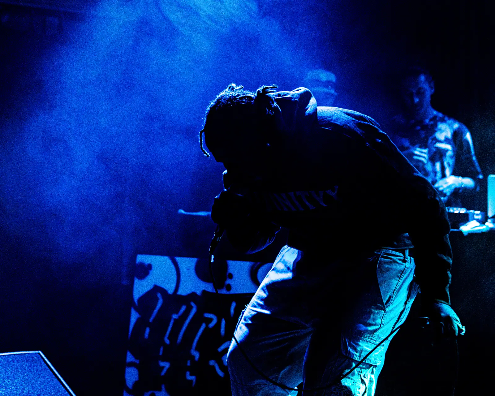 A performer bends forward over the microphone in blue haze, a graffiti-style backdrop behind.