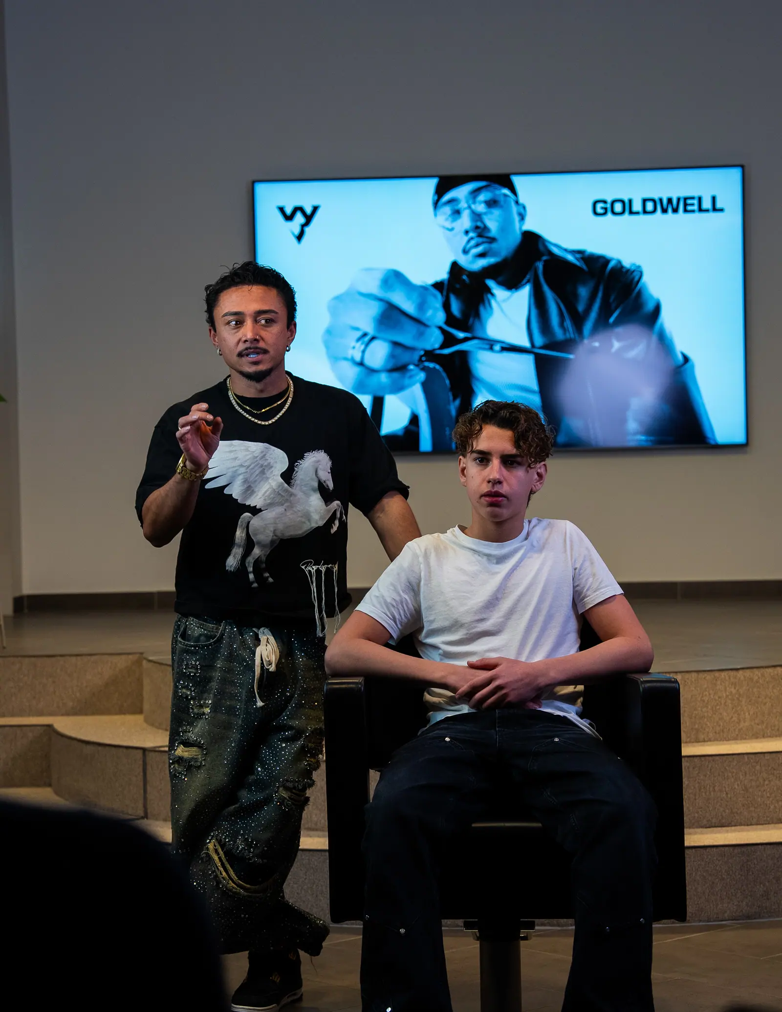 A hairstylist stands beside a young man seated in a chair, a screen showing a portrait behind them.