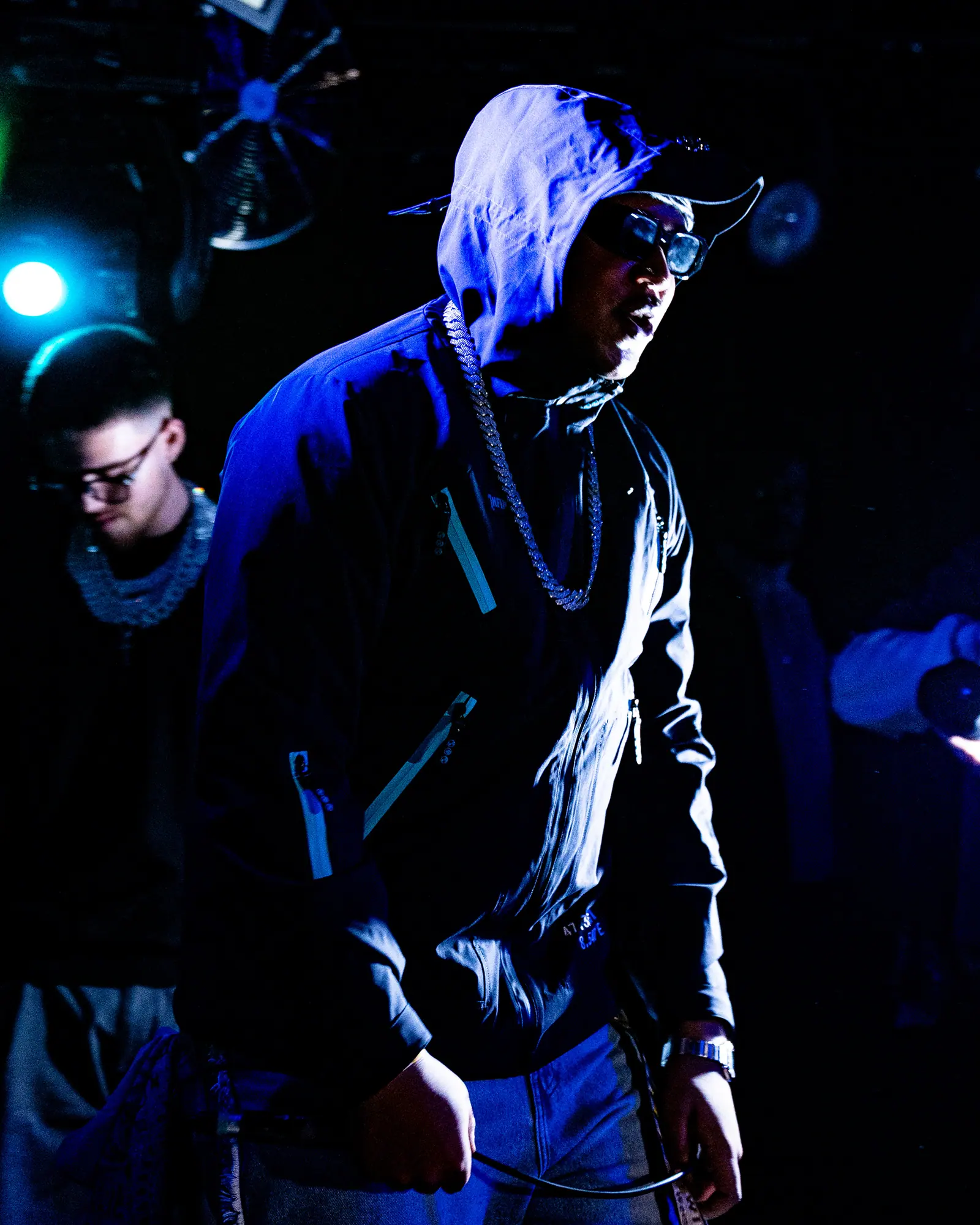 A rapper in a hood, sunglasses and chains looks down from the stage, another performer beside him in blue light.