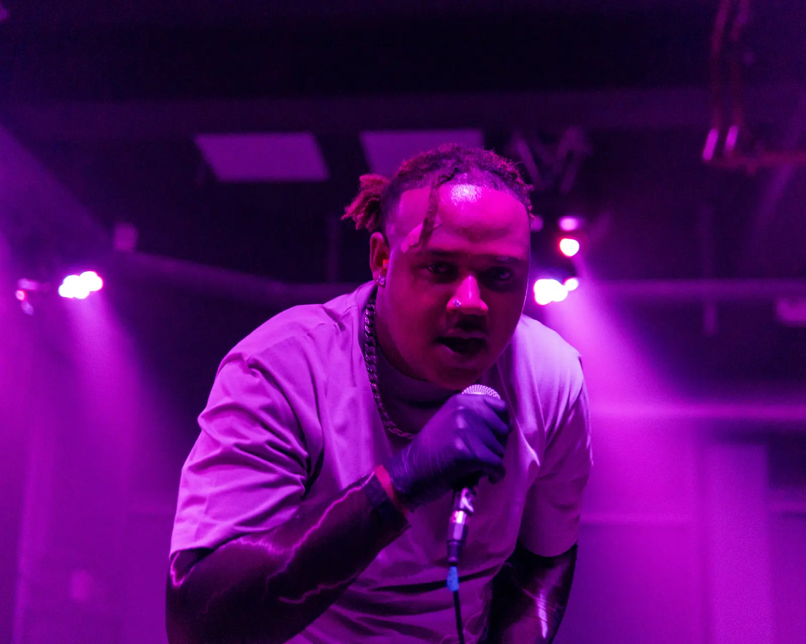 A performer with braided hair sings into a microphone, lit in magenta.