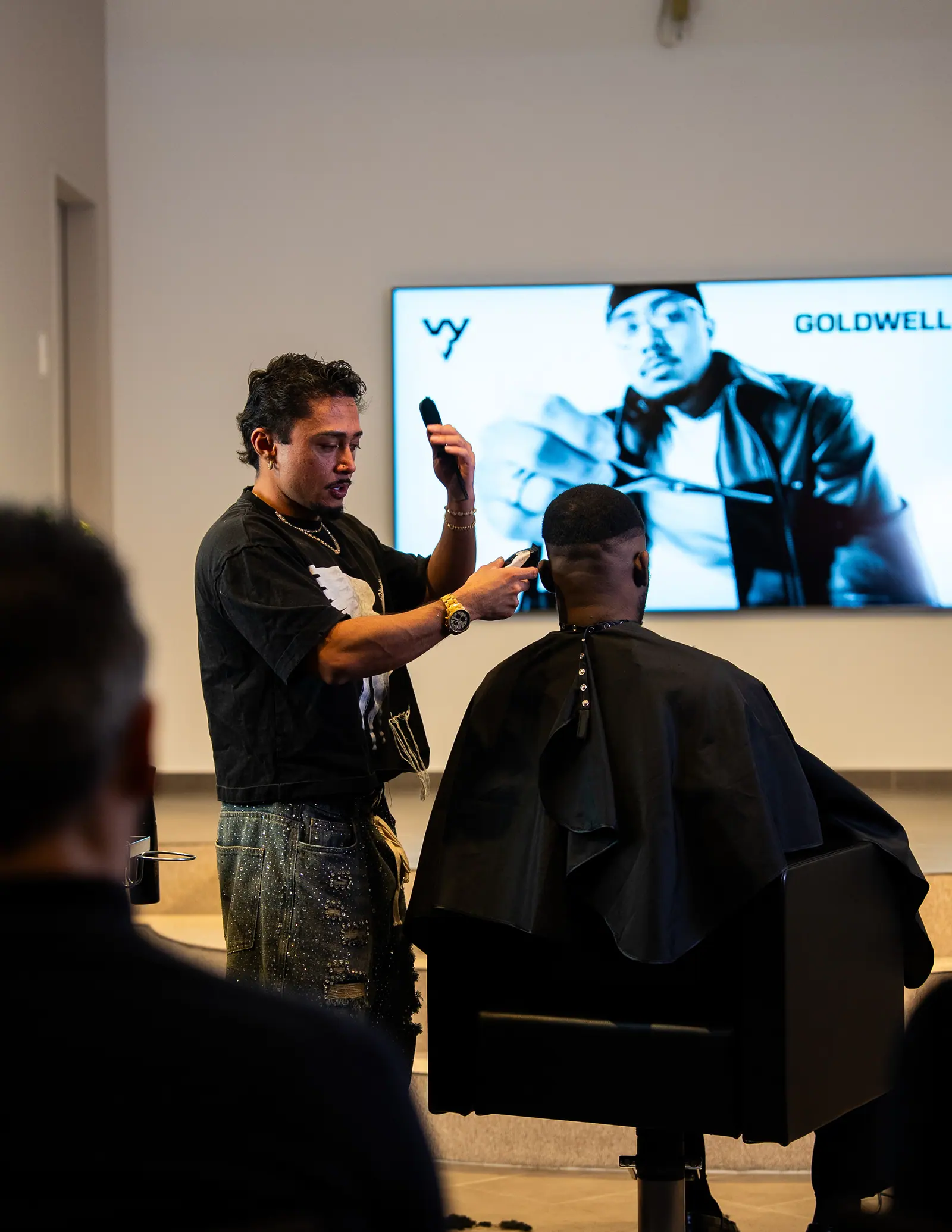 A hairstylist cuts a client’s hair in front of an audience, a screen with a portrait behind them.
