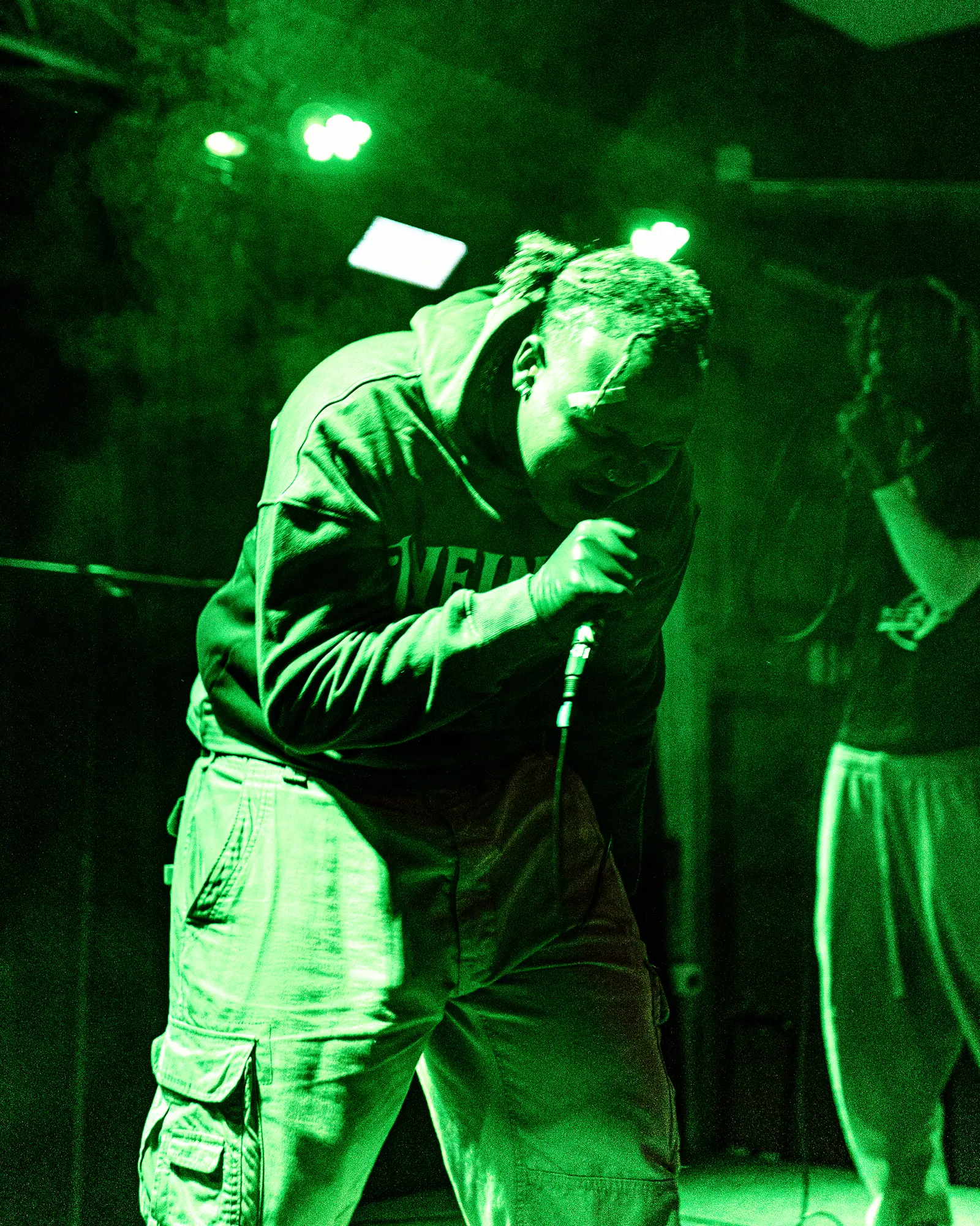 A performer in a hoodie leans into the microphone under green light.