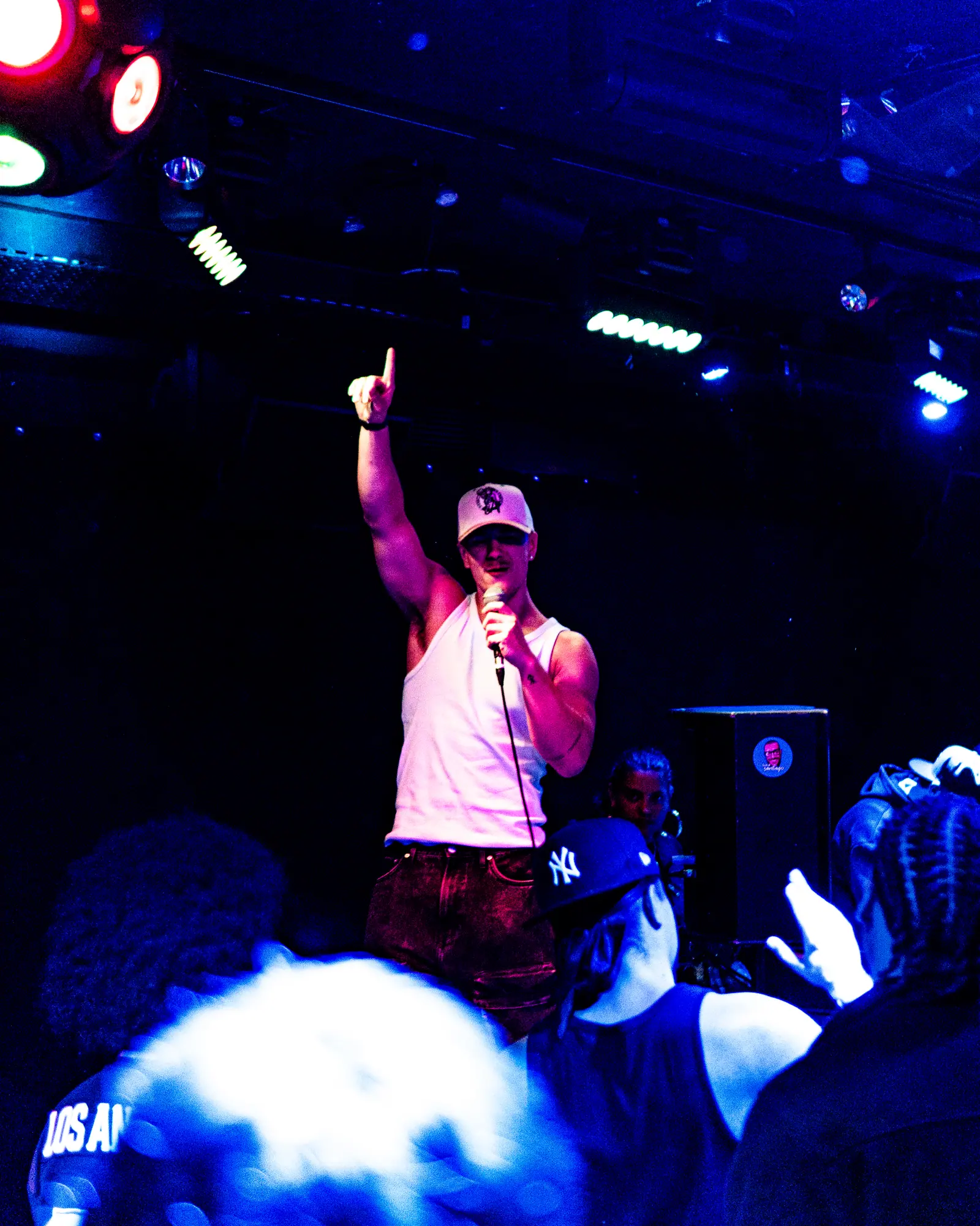 A rapper in a white tank top and cap points up with one hand, microphone in the other, over a crowd lit in deep blue.