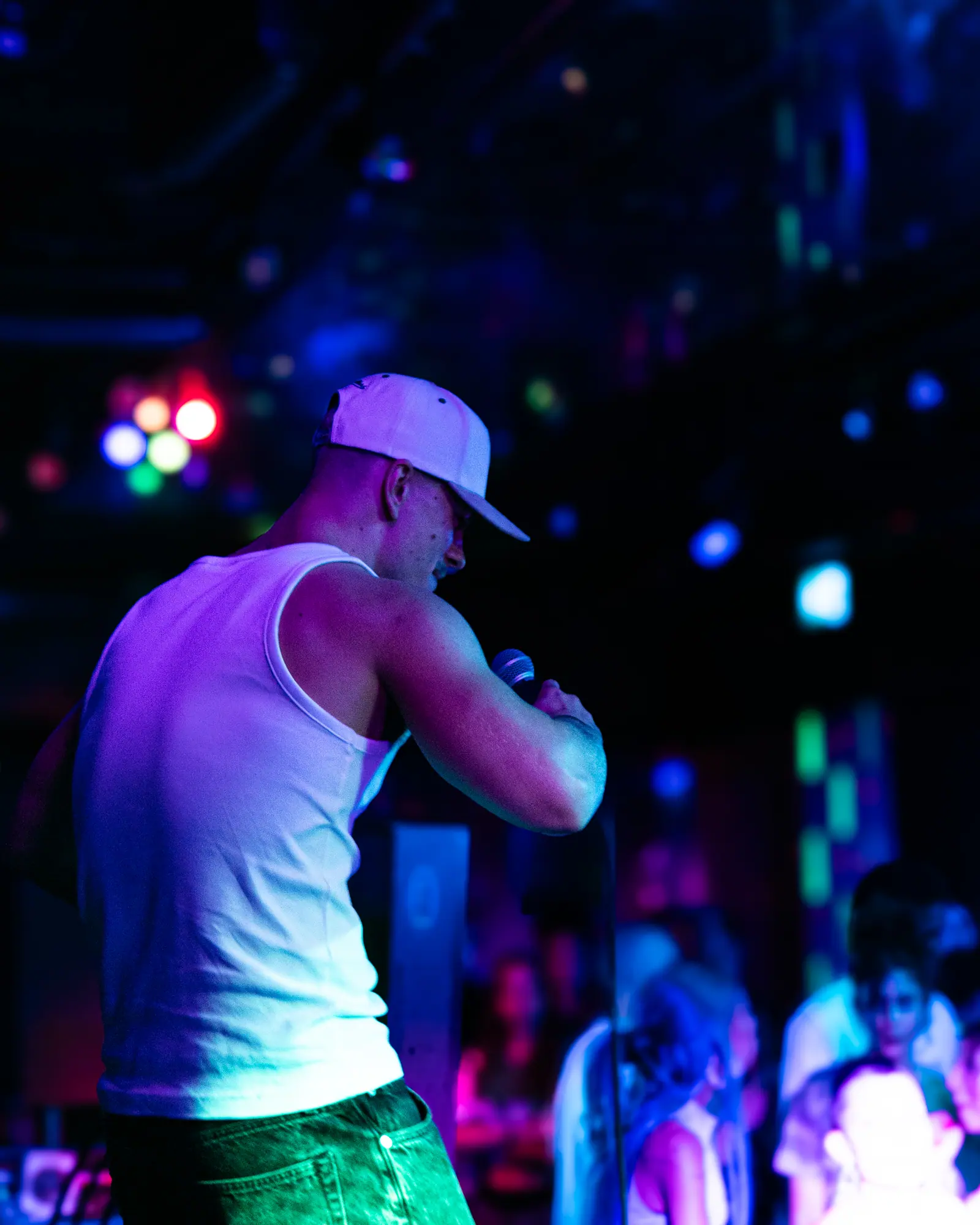 The performer seen in profile, leaning into the microphone, the crowd blurred in pink and violet light behind him.