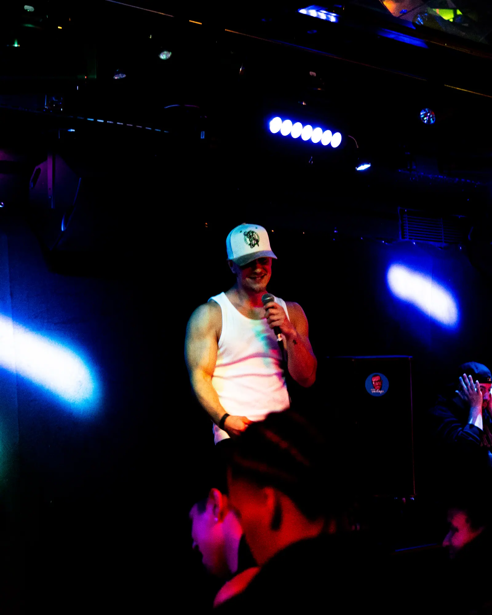 The performer smiles on a dark stage between blue light beams, heads of the crowd in the foreground.