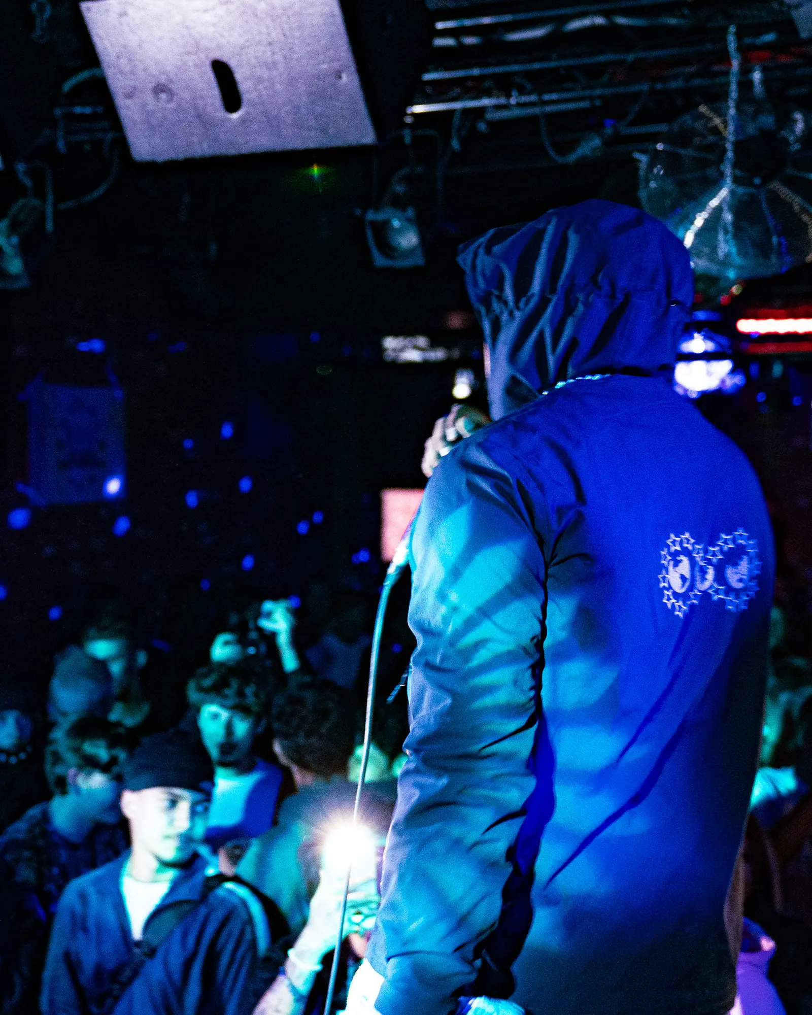 A performer in a hooded jacket seen from behind, facing a crowd holding up their phones.
