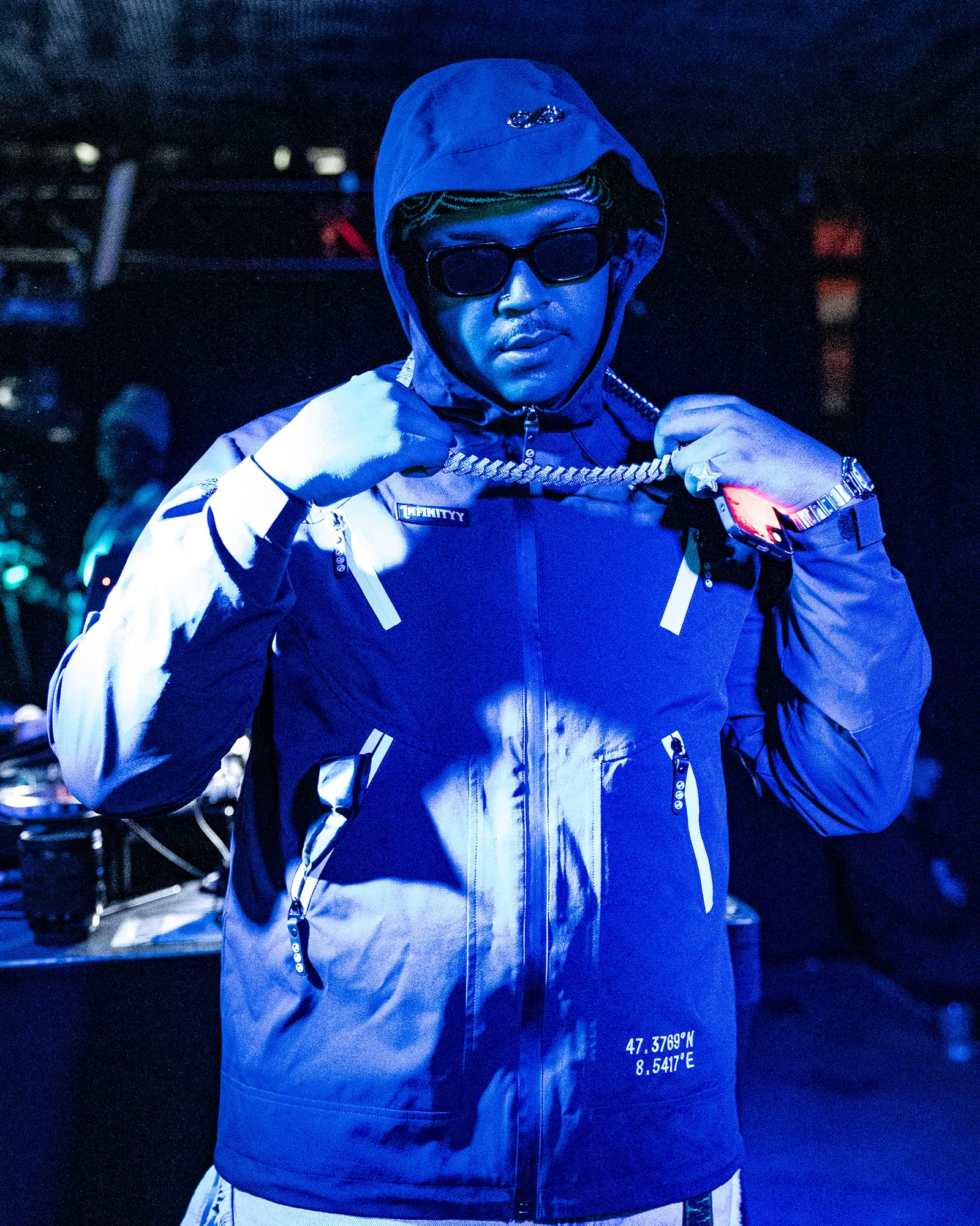 A performer in a hood and sunglasses adjusts the chain around his neck, lit in blue.