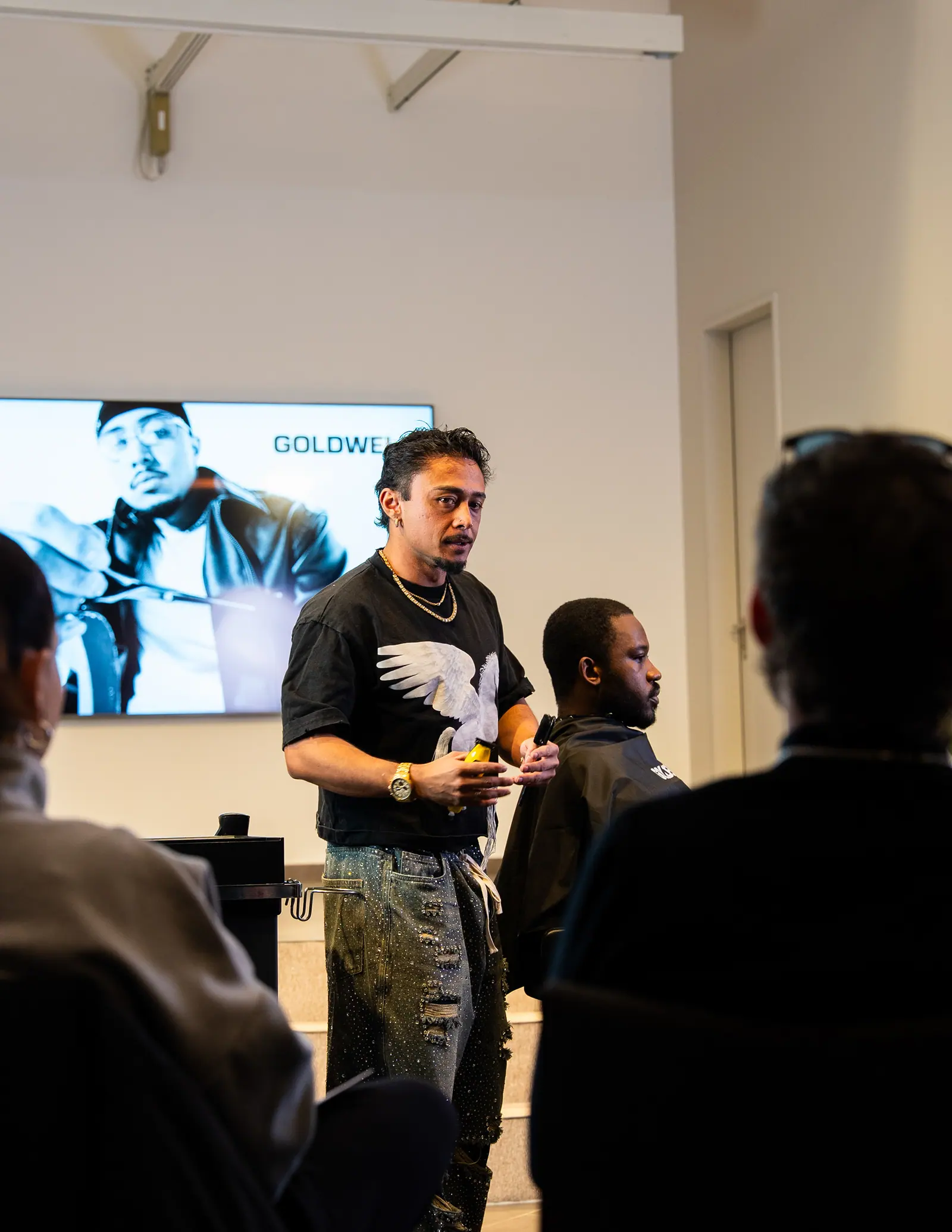 A man in a printed T-shirt speaks to a seated audience in a bright room, a screen with a portrait behind him.