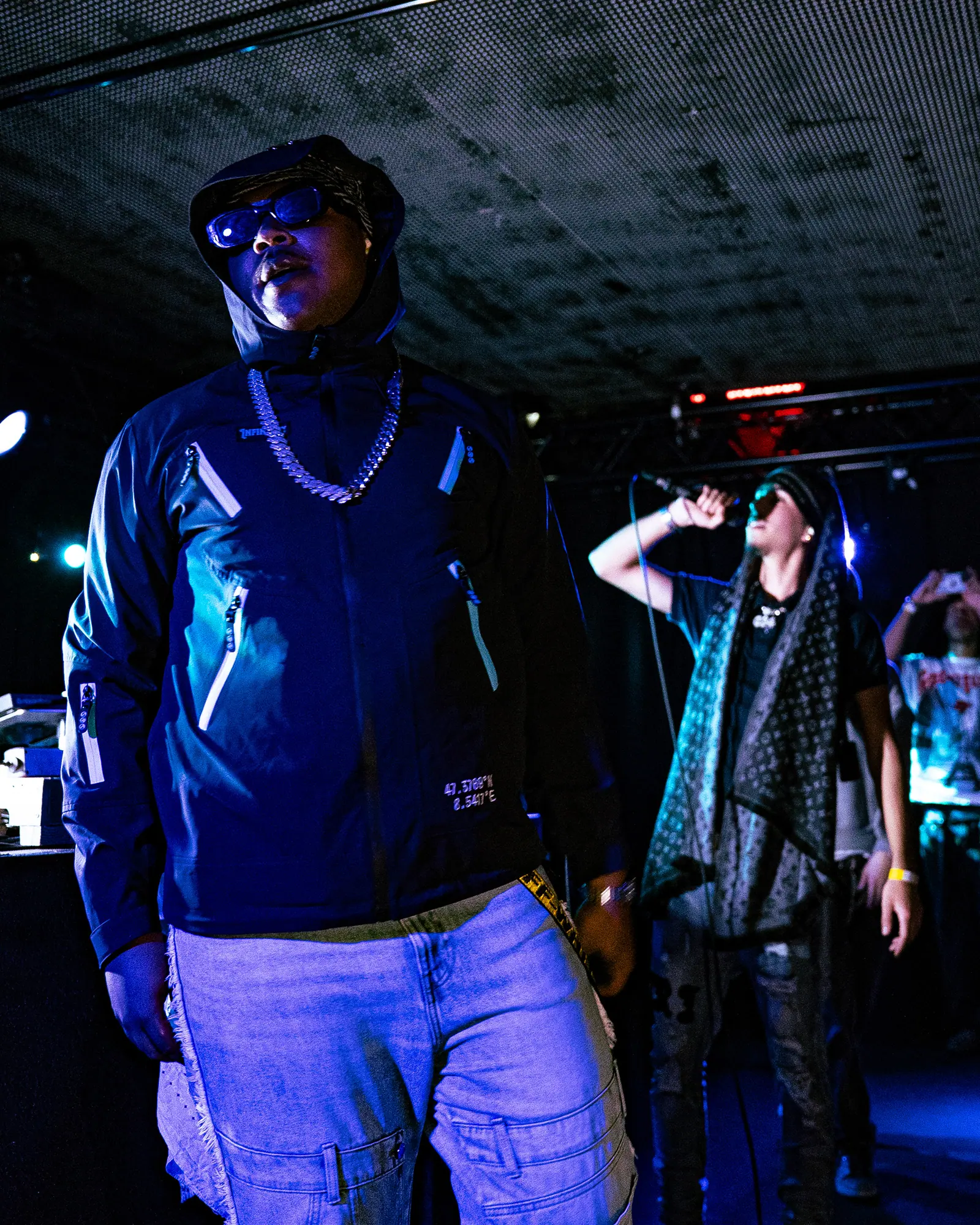 A performer in sunglasses, chains and a zipped jacket stands on stage, another artist singing behind him.