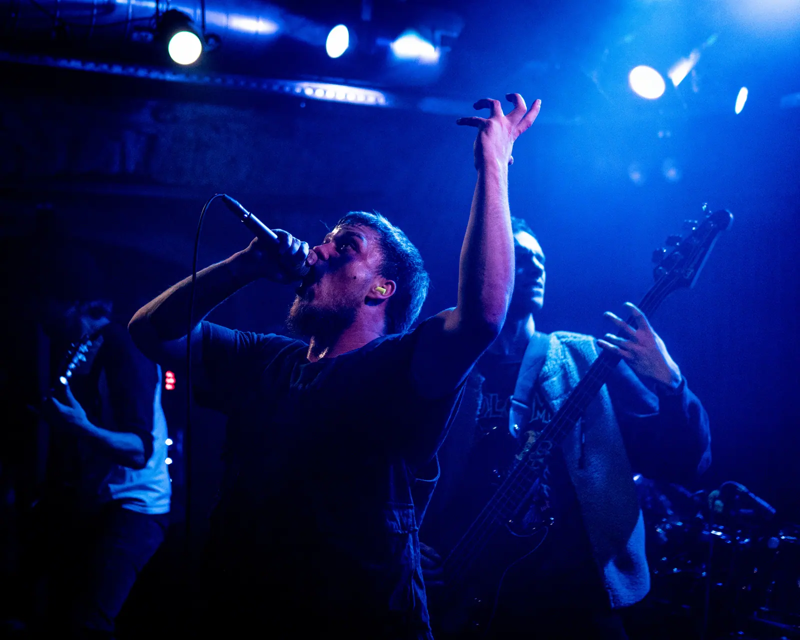 A singer holds the microphone up as he performs with a band in deep blue light.