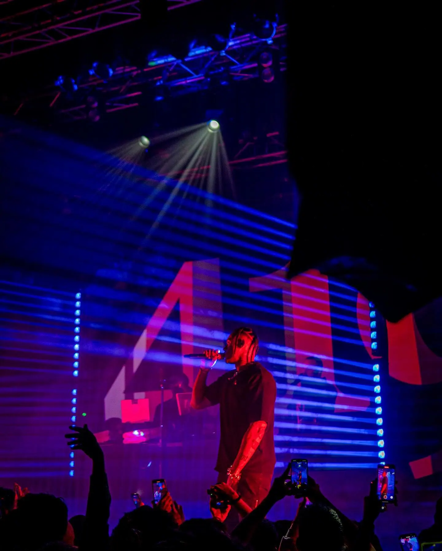 A rapper with braids performs under purple and blue stage light, large illuminated numbers behind him, phones raised in the crowd below.