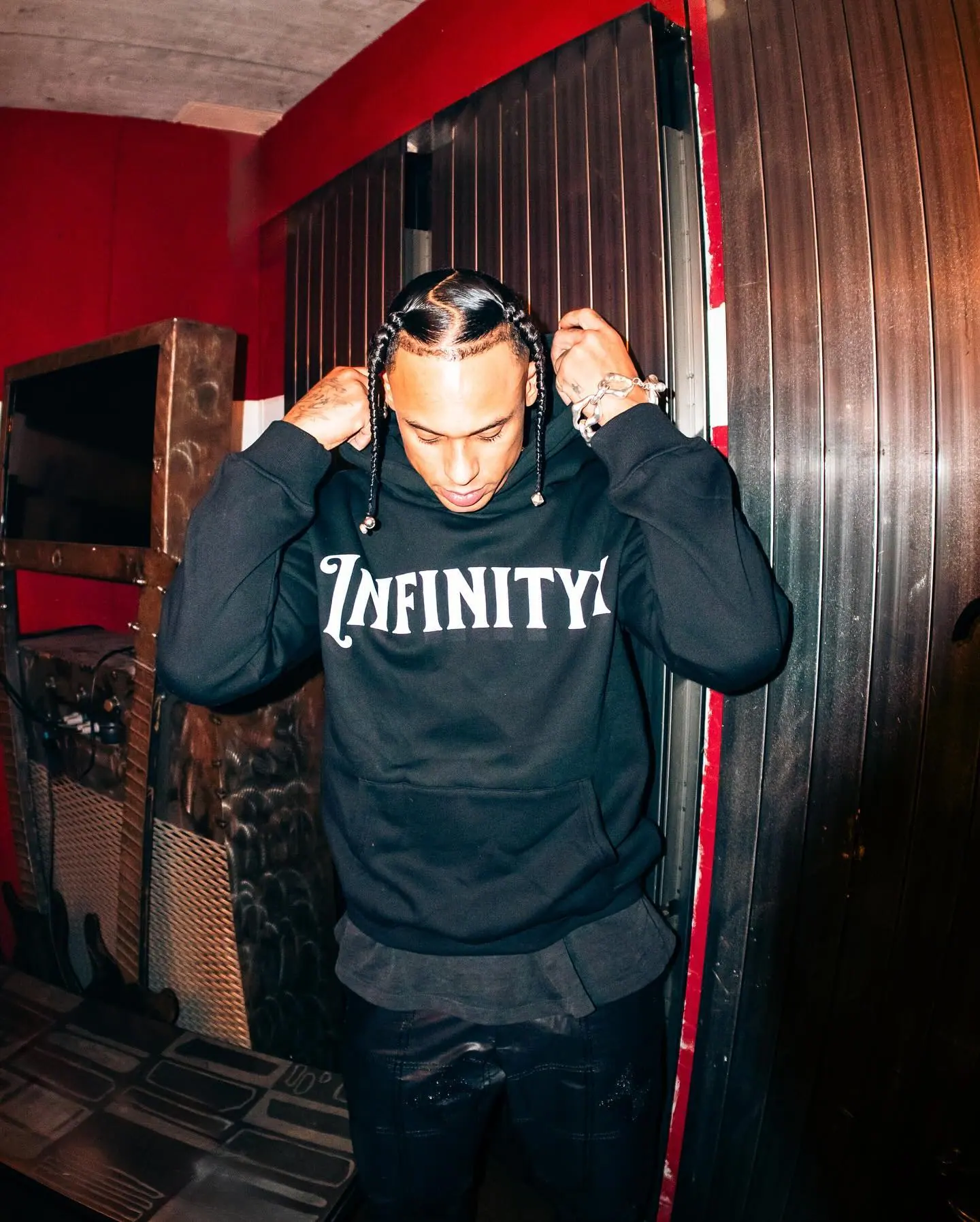 Backstage, a man with braids pulls up the hood of a black Infinityy hoodie in front of a red wall and dark wooden panels.