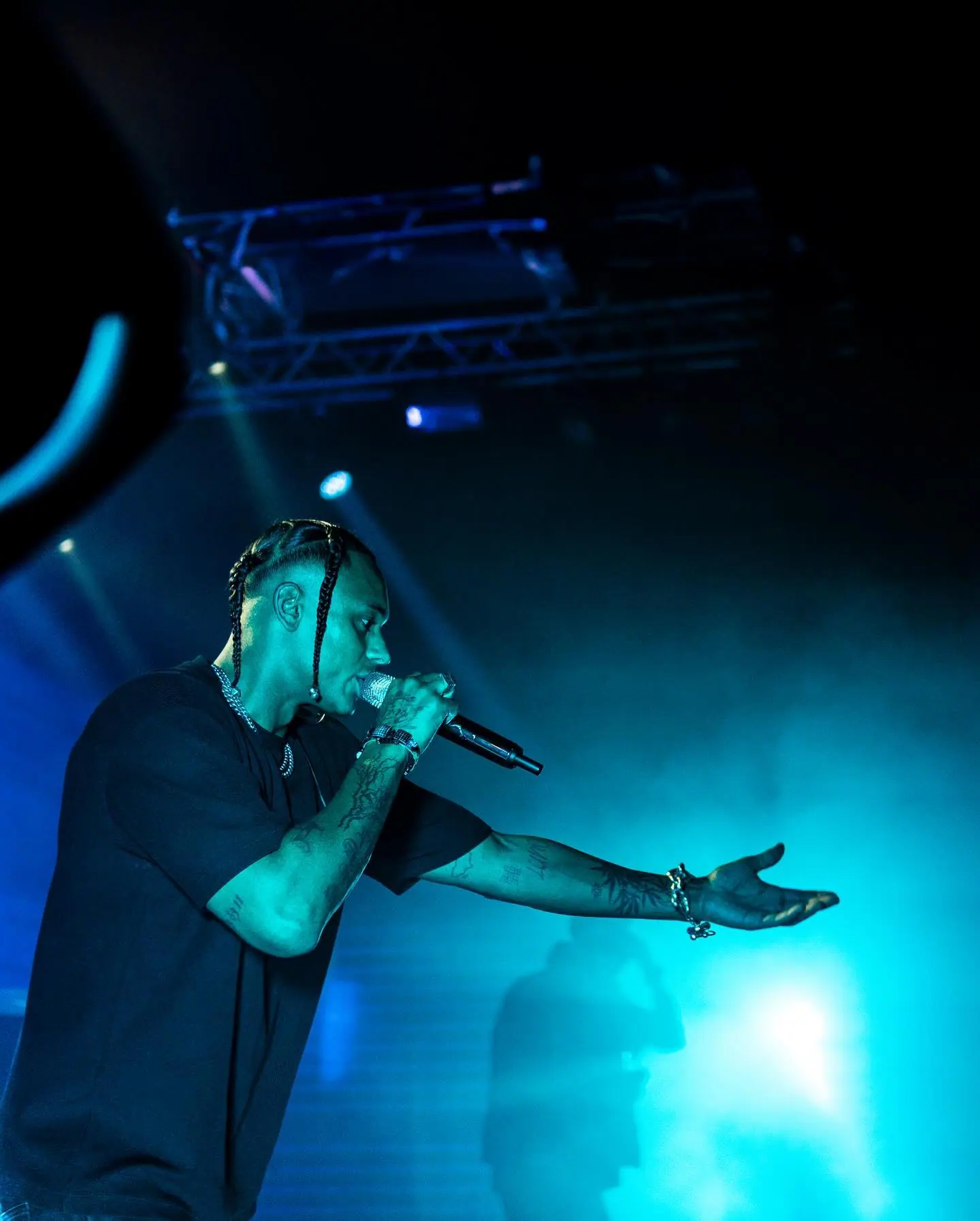 A rapper in a black T-shirt raps into a microphone and reaches out toward the crowd through blue haze and stage light.