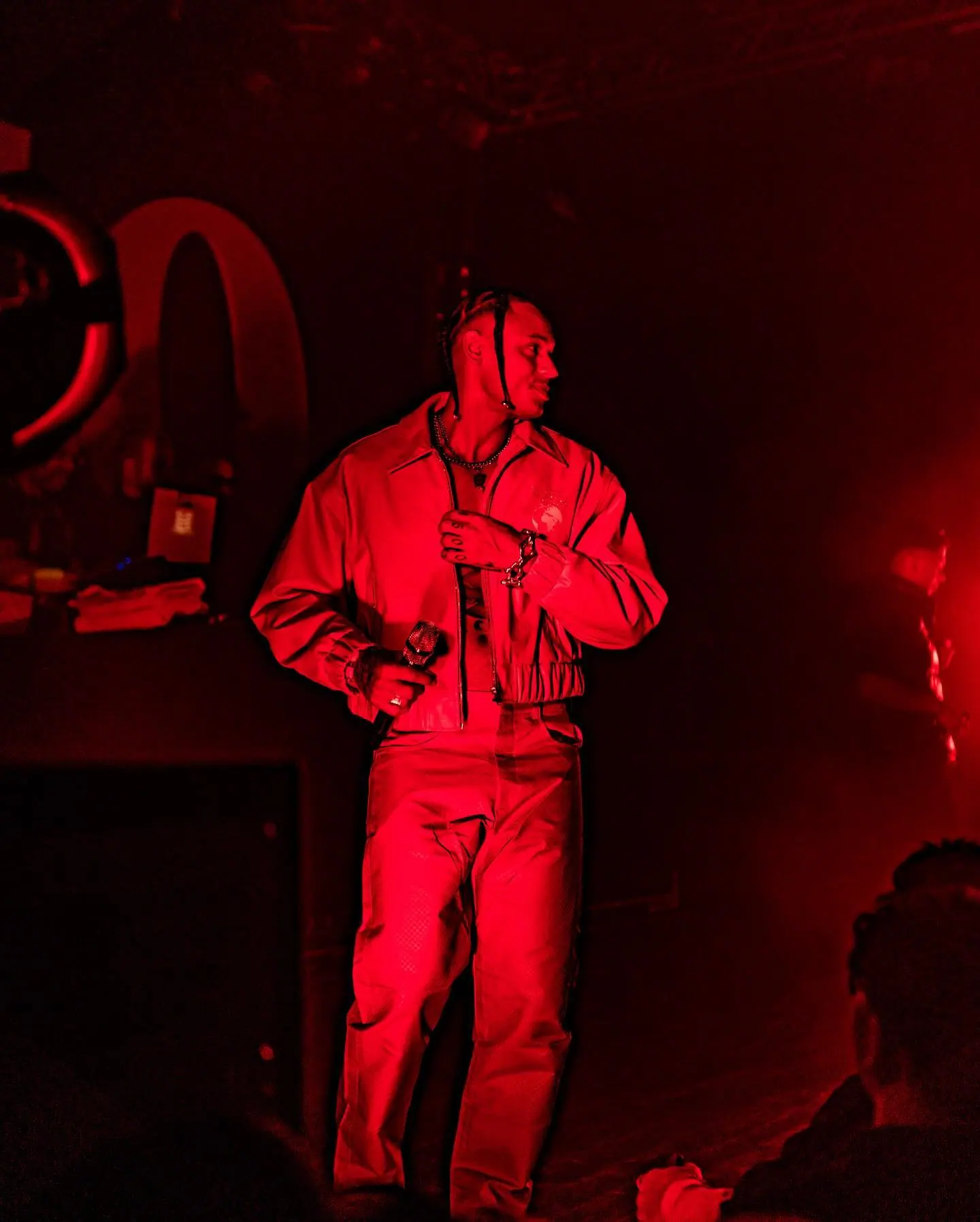 A performer in a red jacket and trousers stands on stage in deep red light, microphone in hand.