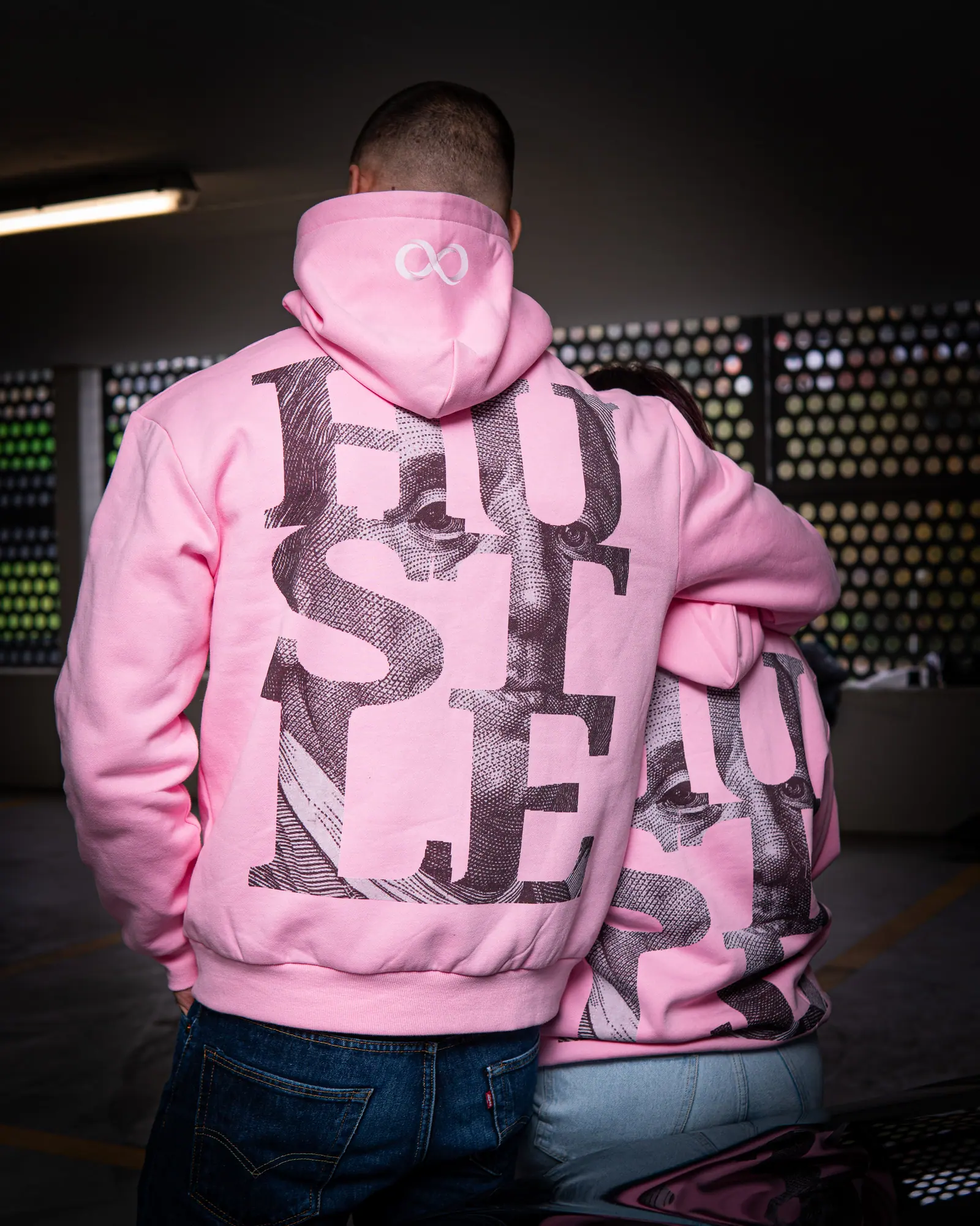 A man seen from behind in a pink hoodie with an oversized HUSTLE back print, a second person leaning into his shoulder.