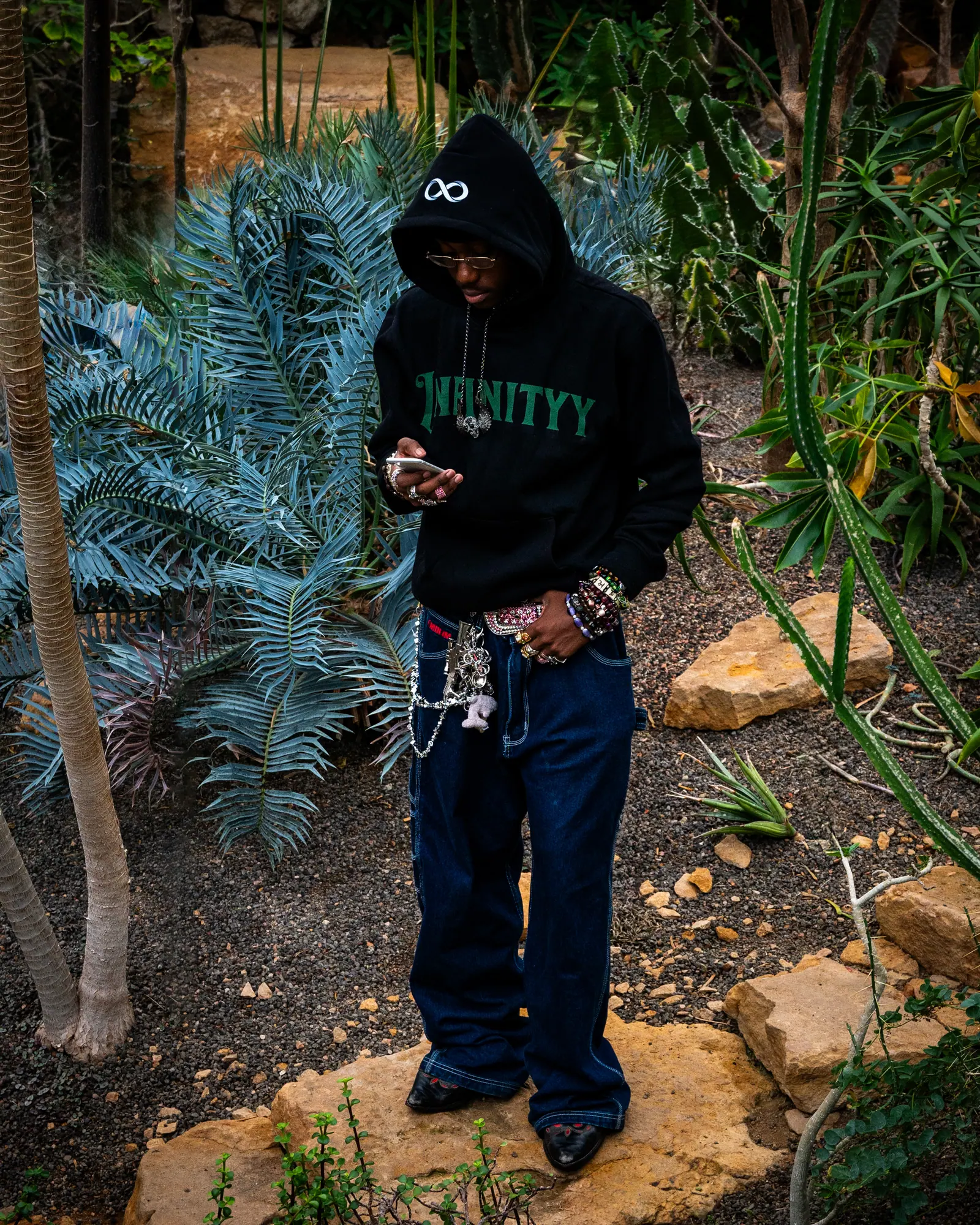 A man in a black Infinityy hoodie and baggy jeans stands among ferns and rocks in a greenhouse, looking down at his phone.
