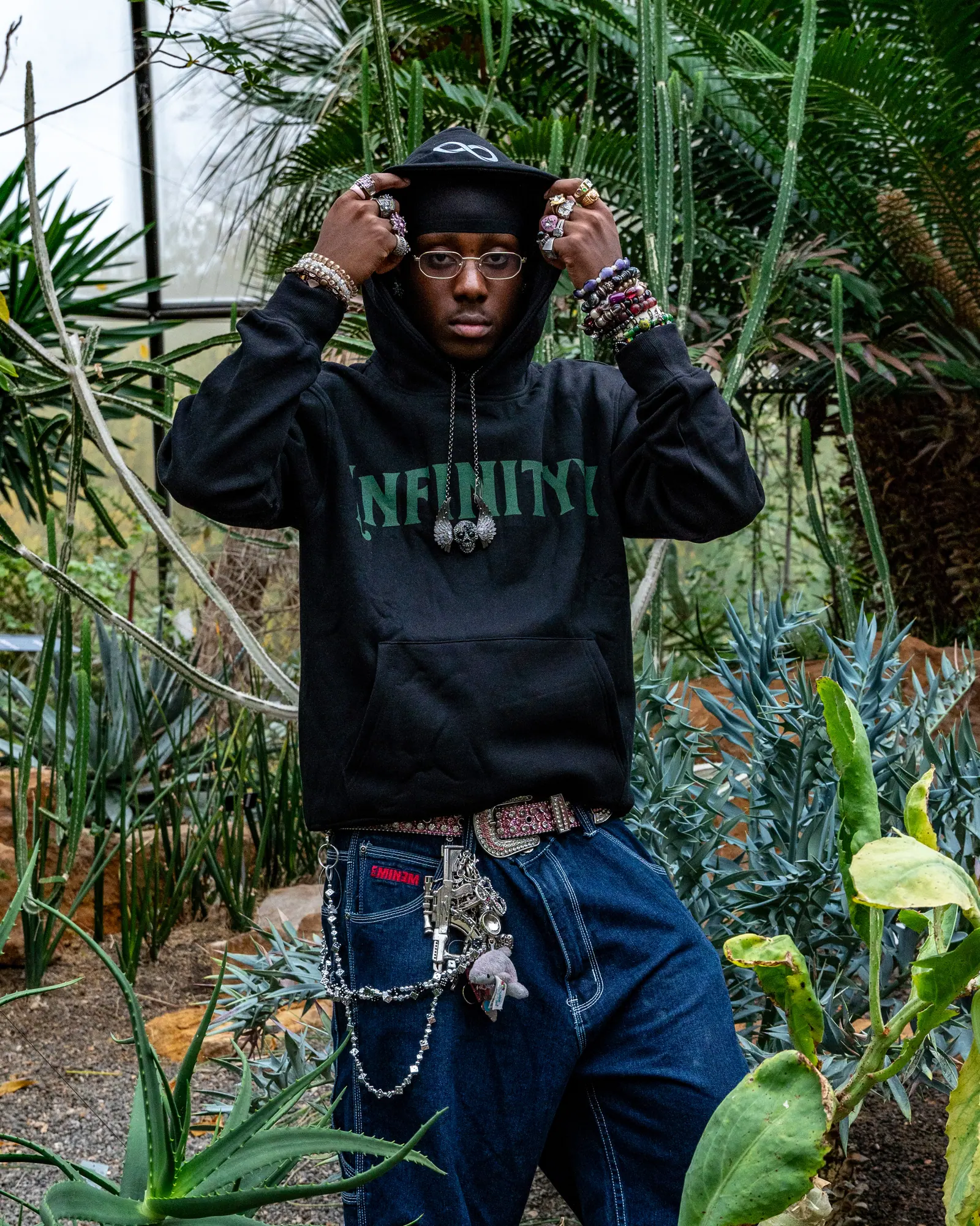 A man in a black Infinityy hoodie and beanie holds his hood with both hands, silver rings and chains, tropical plants behind him.
