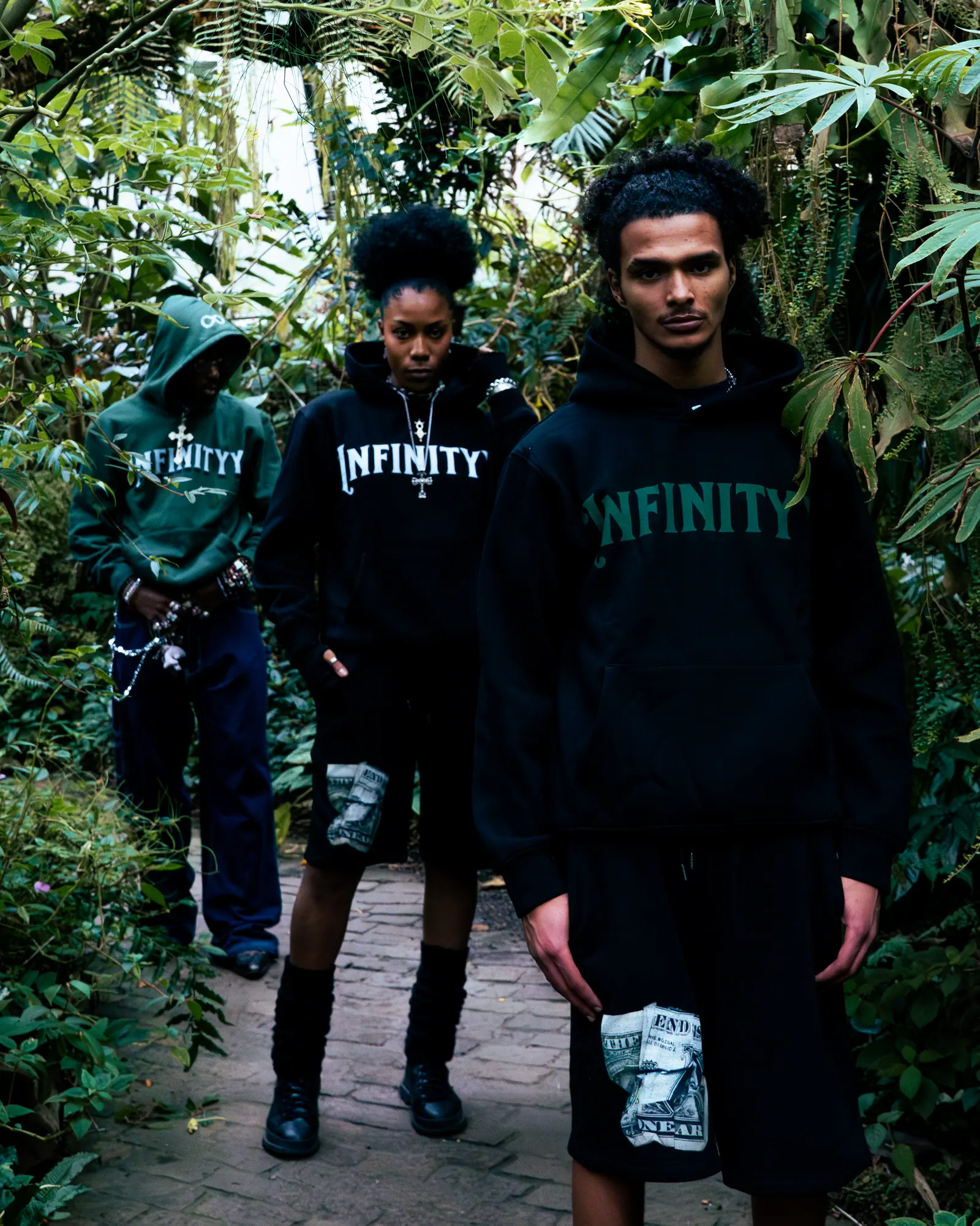 Three models in black and green Infinityy hoodies walk toward the camera along a greenhouse path.
