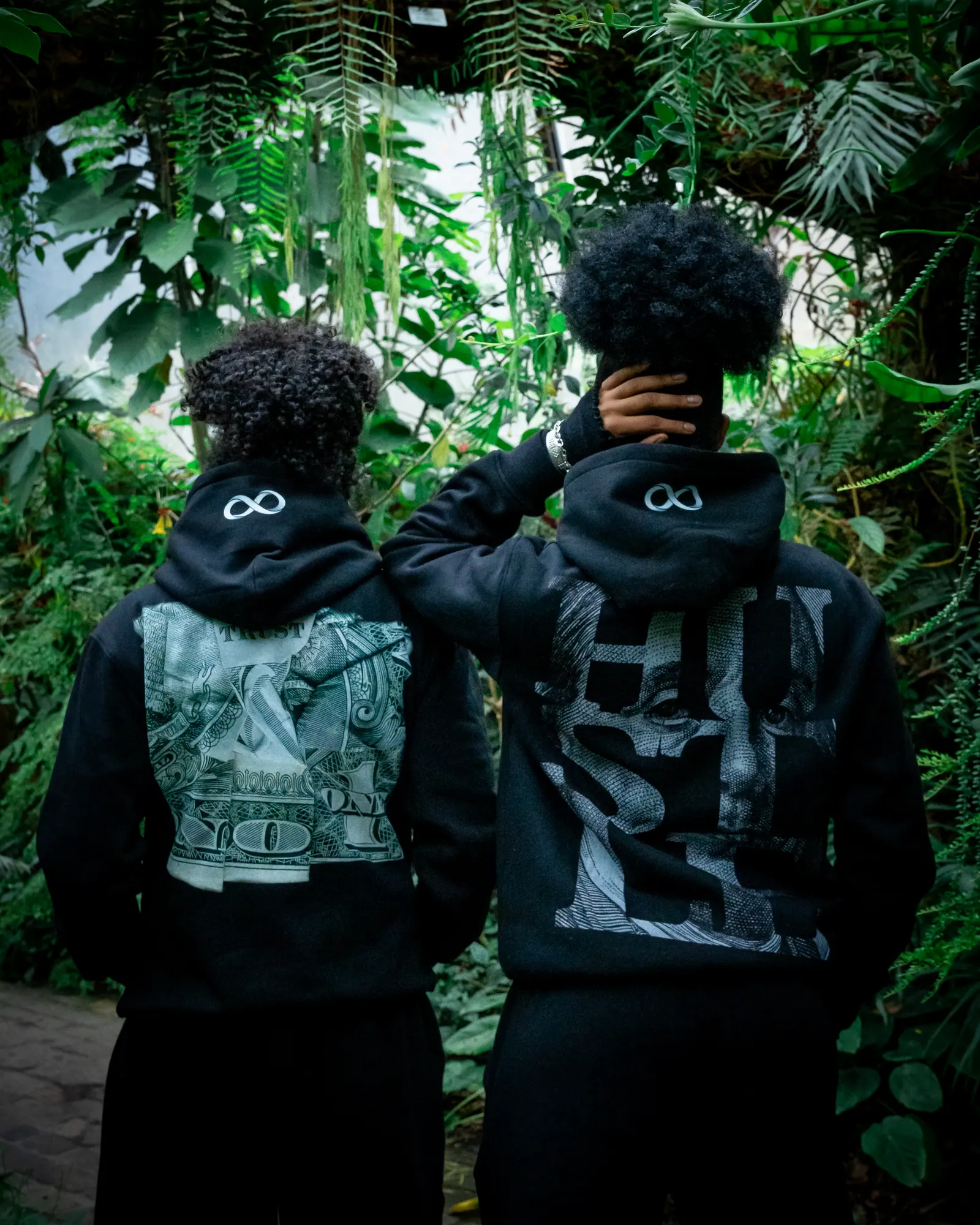 Two people seen from behind in black hoodies, one with a dollar-bill back print and one with the HUSTLE print, framed by dense leaves.