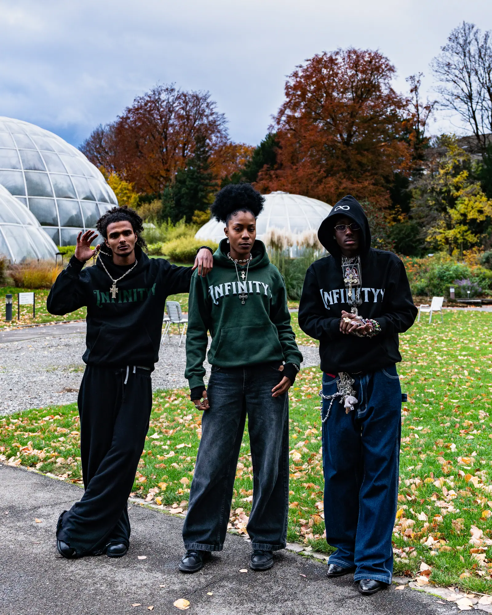 Three models in black and green hoodies stand on a gravel path in a botanical garden, glass domes and autumn trees behind them.
