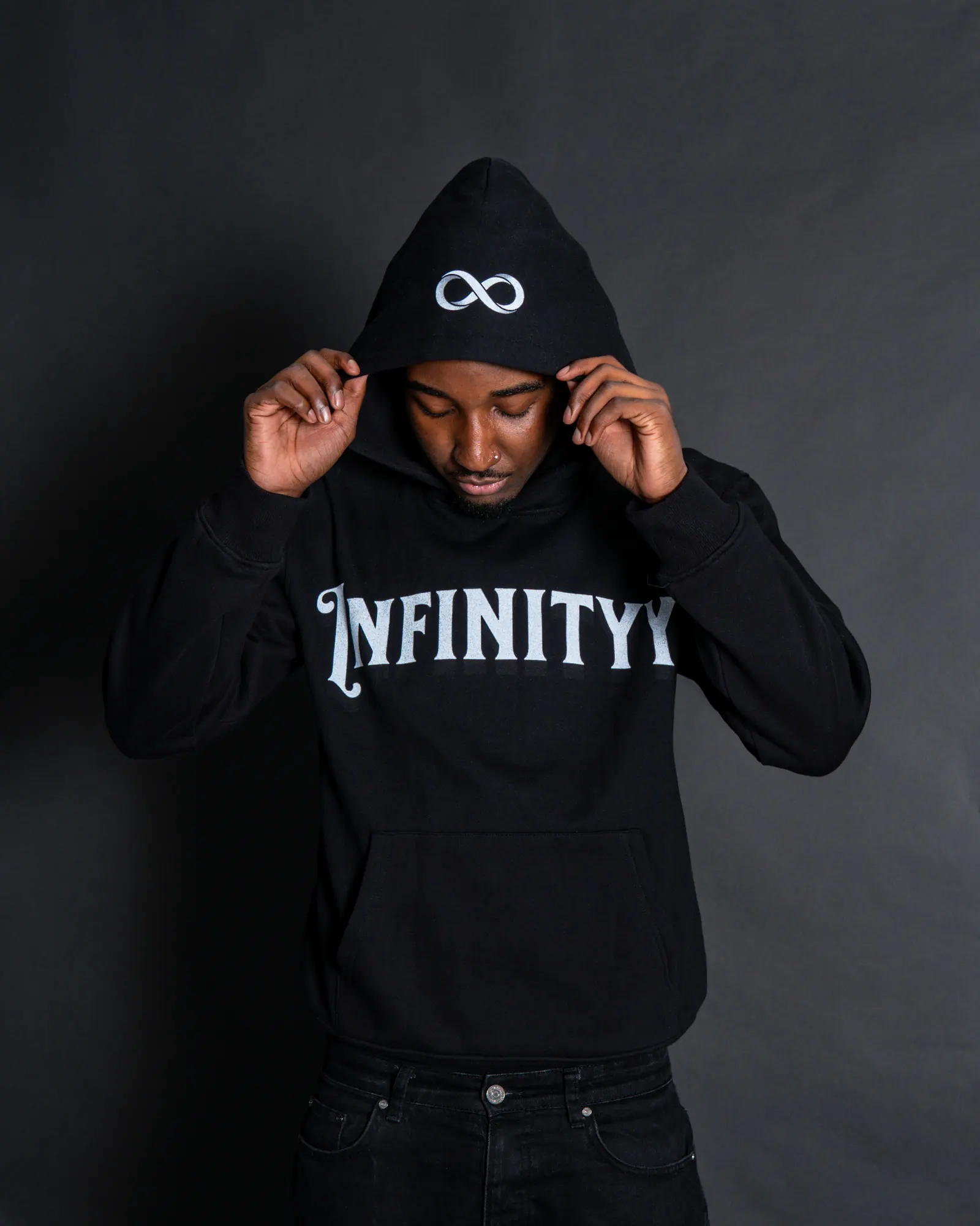 A man pulls up the hood of a black Infinityy hoodie against a dark grey studio backdrop.