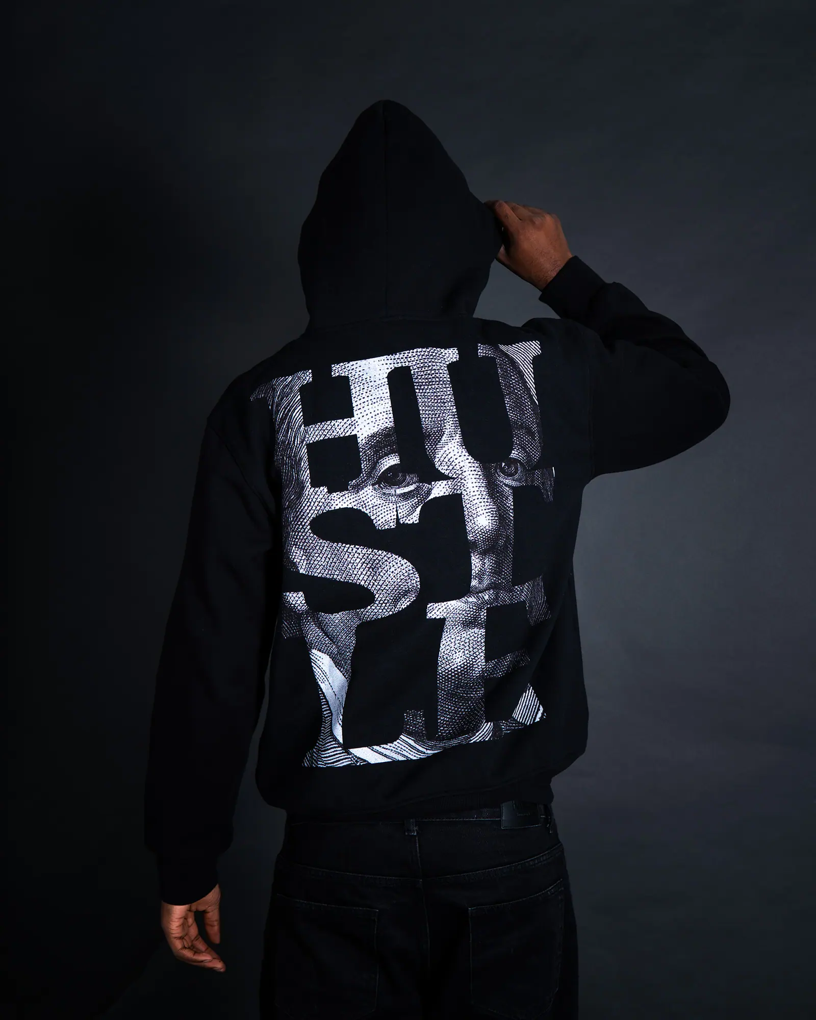 A man seen from behind in a black hoodie with a silver HUSTLE back print, one hand on his hood.