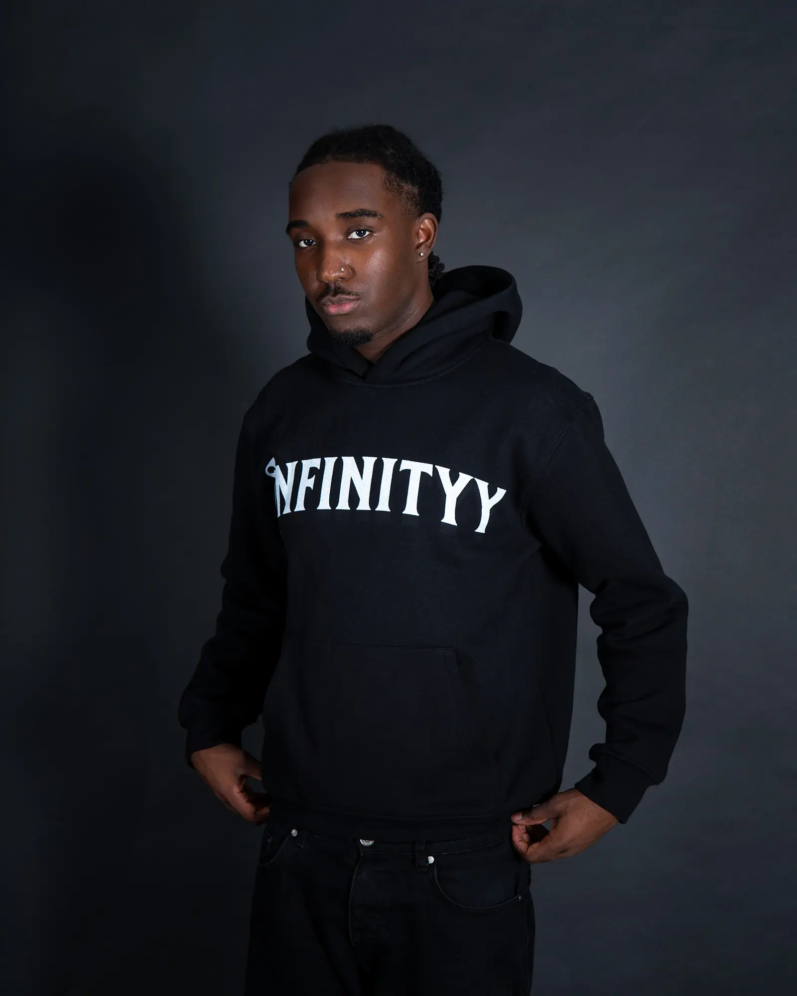 A man in a black Infinityy hoodie stands with his arms relaxed against a dark studio backdrop.