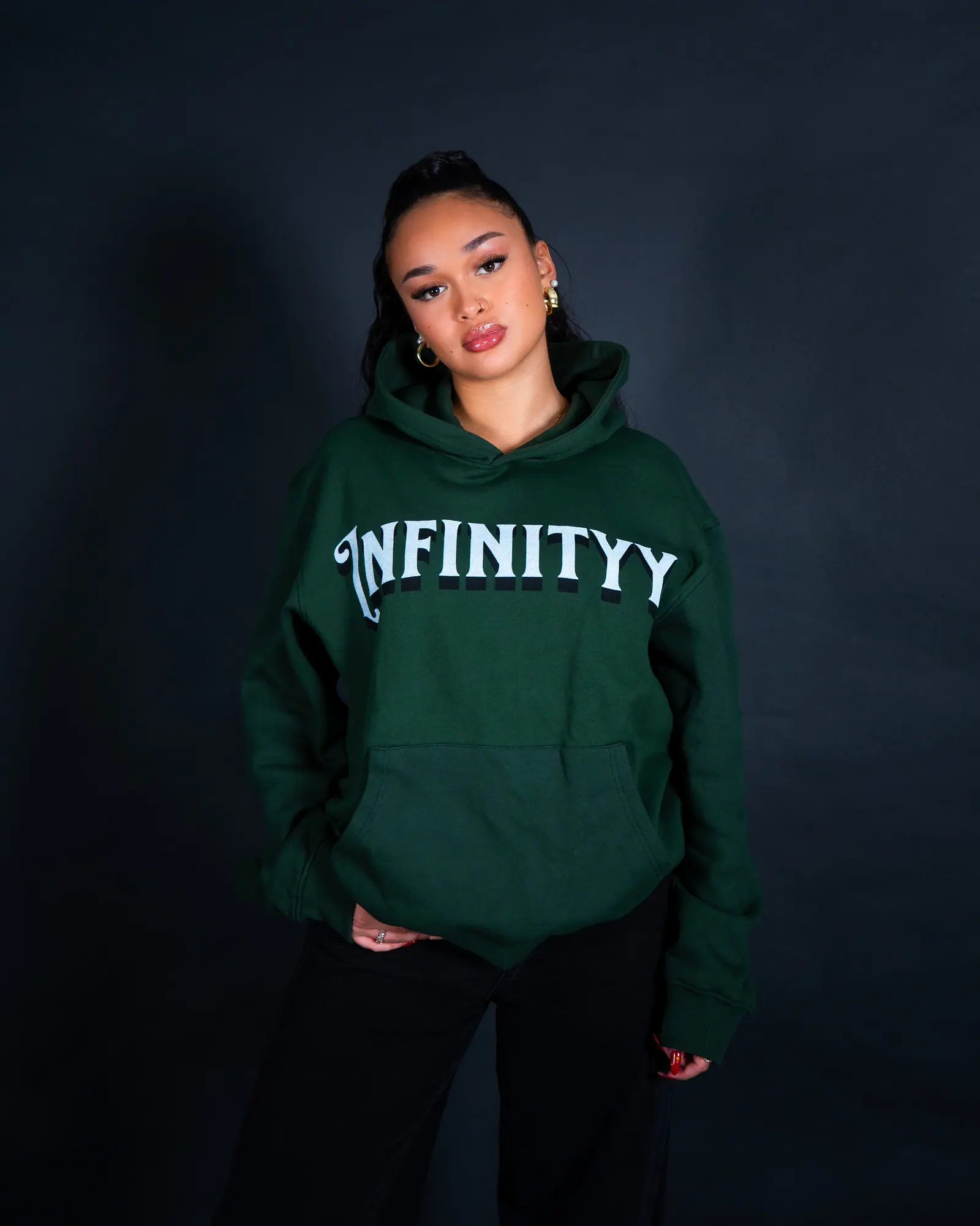 A woman in a green Infinityy hoodie looks straight into the camera with her hands in her pockets.