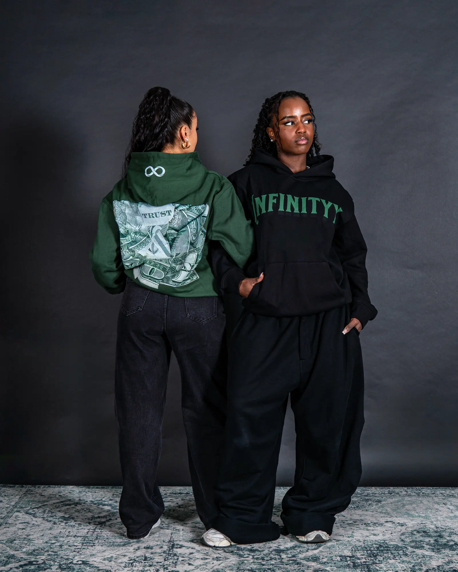 Two women in the studio: one turned away to show the dollar-bill print on a green hoodie, the other facing forward in black.