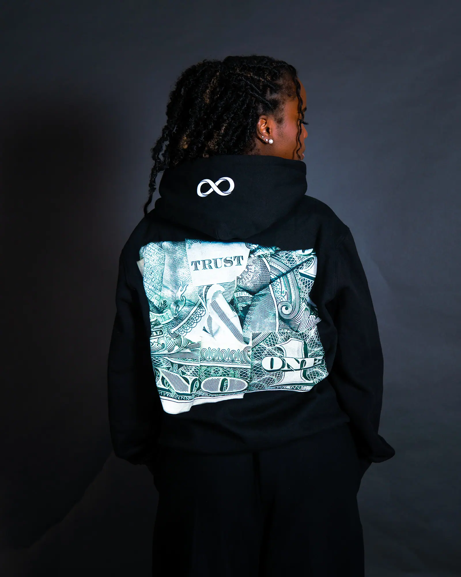 A woman seen from behind in a black hoodie with a large dollar-bill back print and an infinity symbol on the hood.