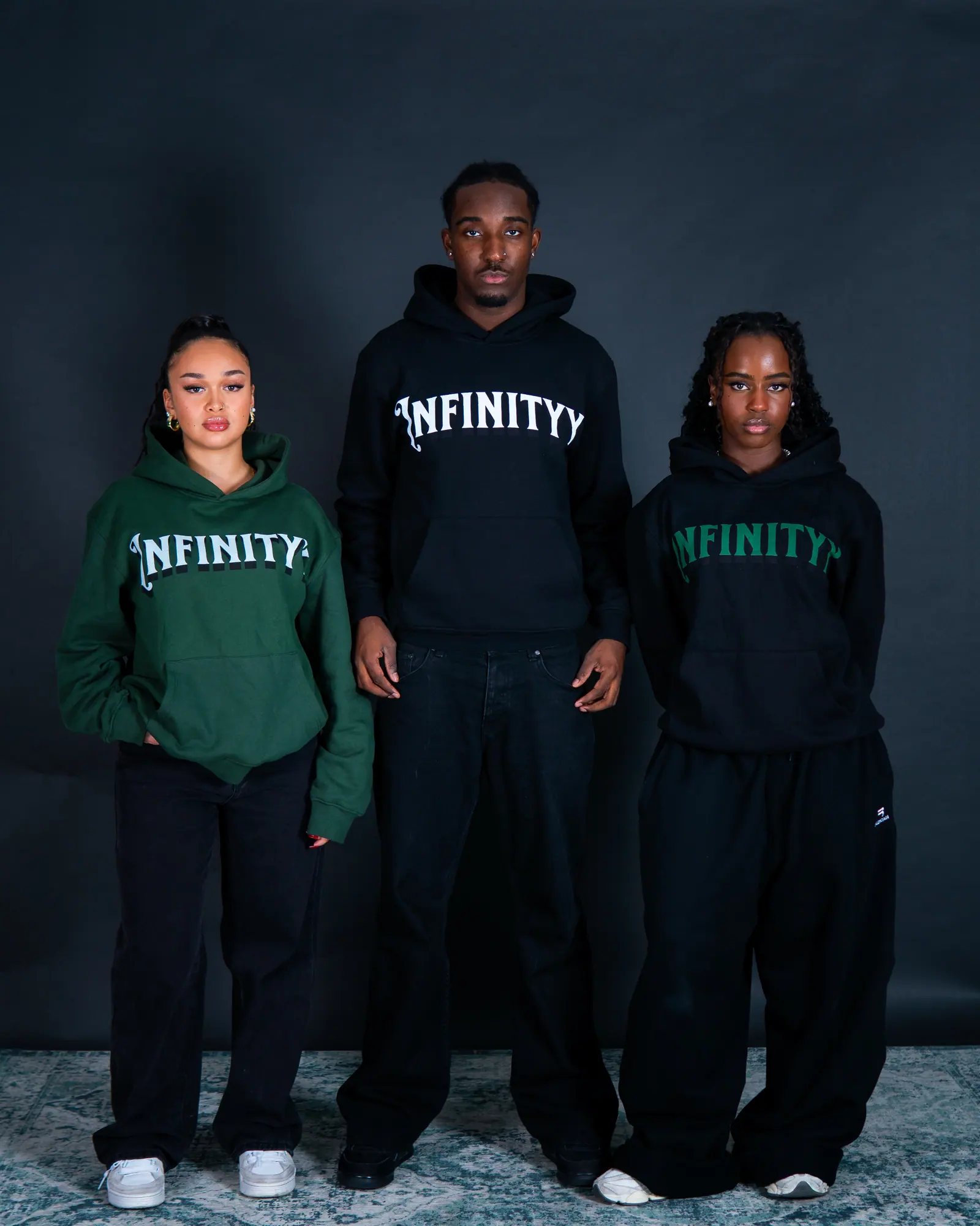 Three models in green and black Infinityy hoodies and wide black trousers stand together against the studio backdrop.