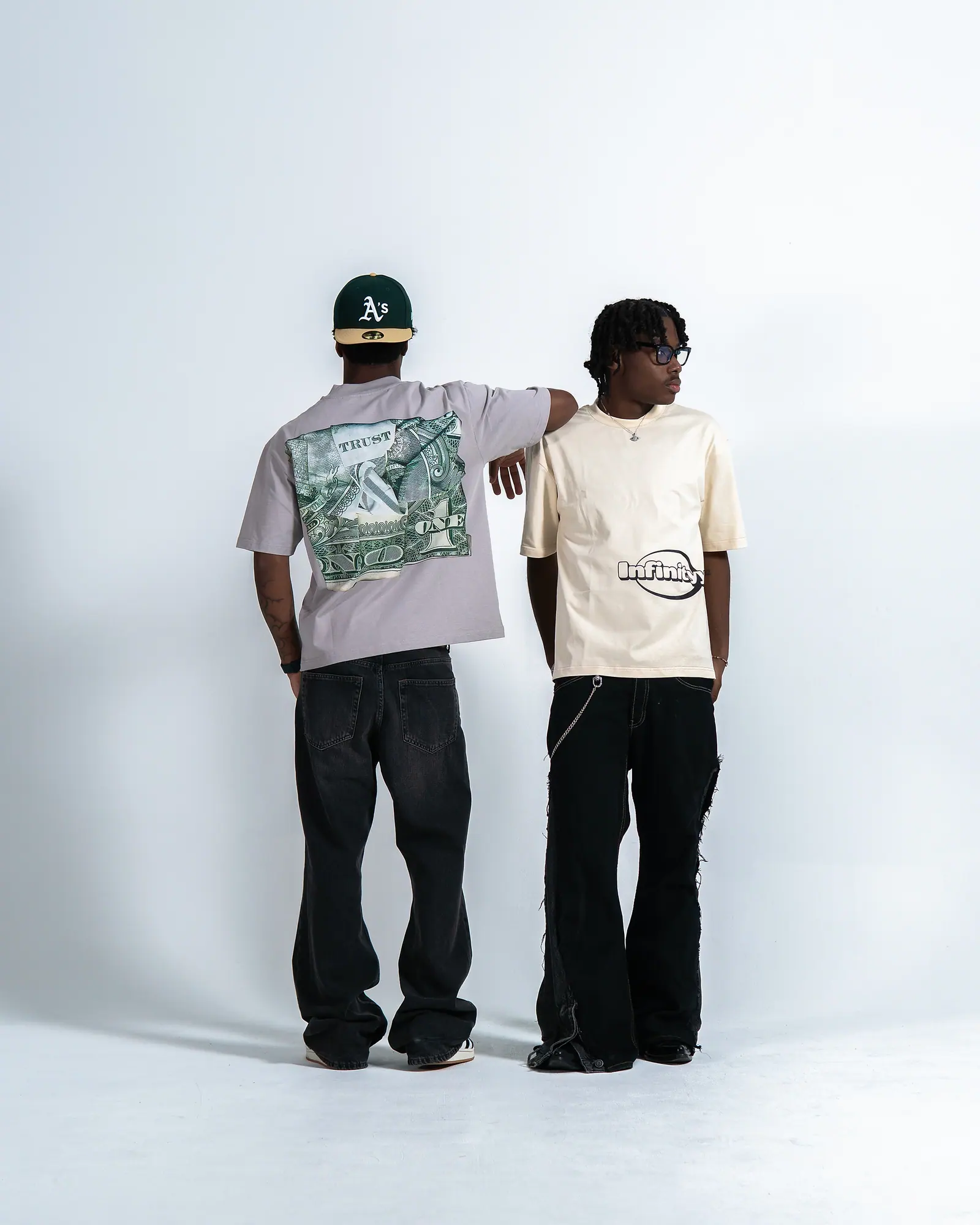 Two men against a white studio wall: one turned away in a grey T-shirt with a dollar-bill back print and a green cap, the other facing sideways in a cream Infinityy T-shirt.