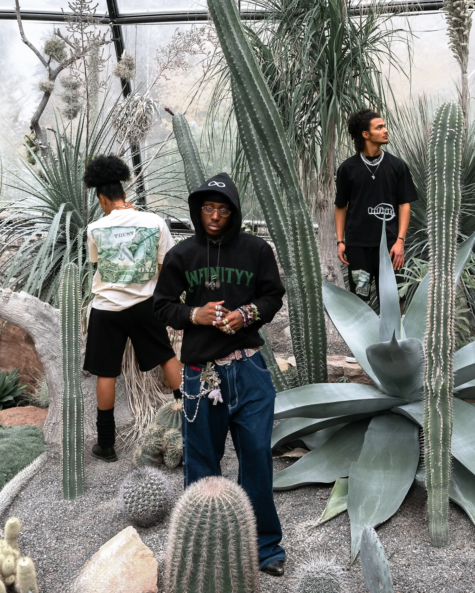 Three models among tall cacti and agaves in a greenhouse, the one in front in a black Infinityy hoodie with chains and wide jeans.