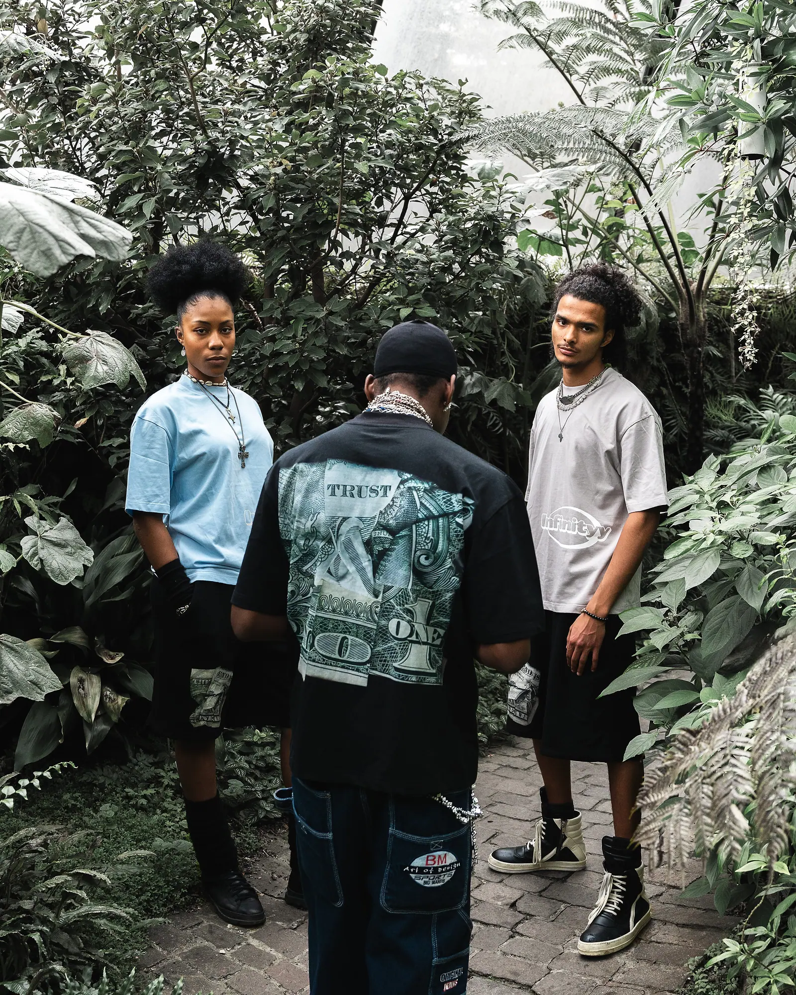 Three models in a leafy greenhouse, the one in the middle turned away to show a dollar-bill print on a black T-shirt.