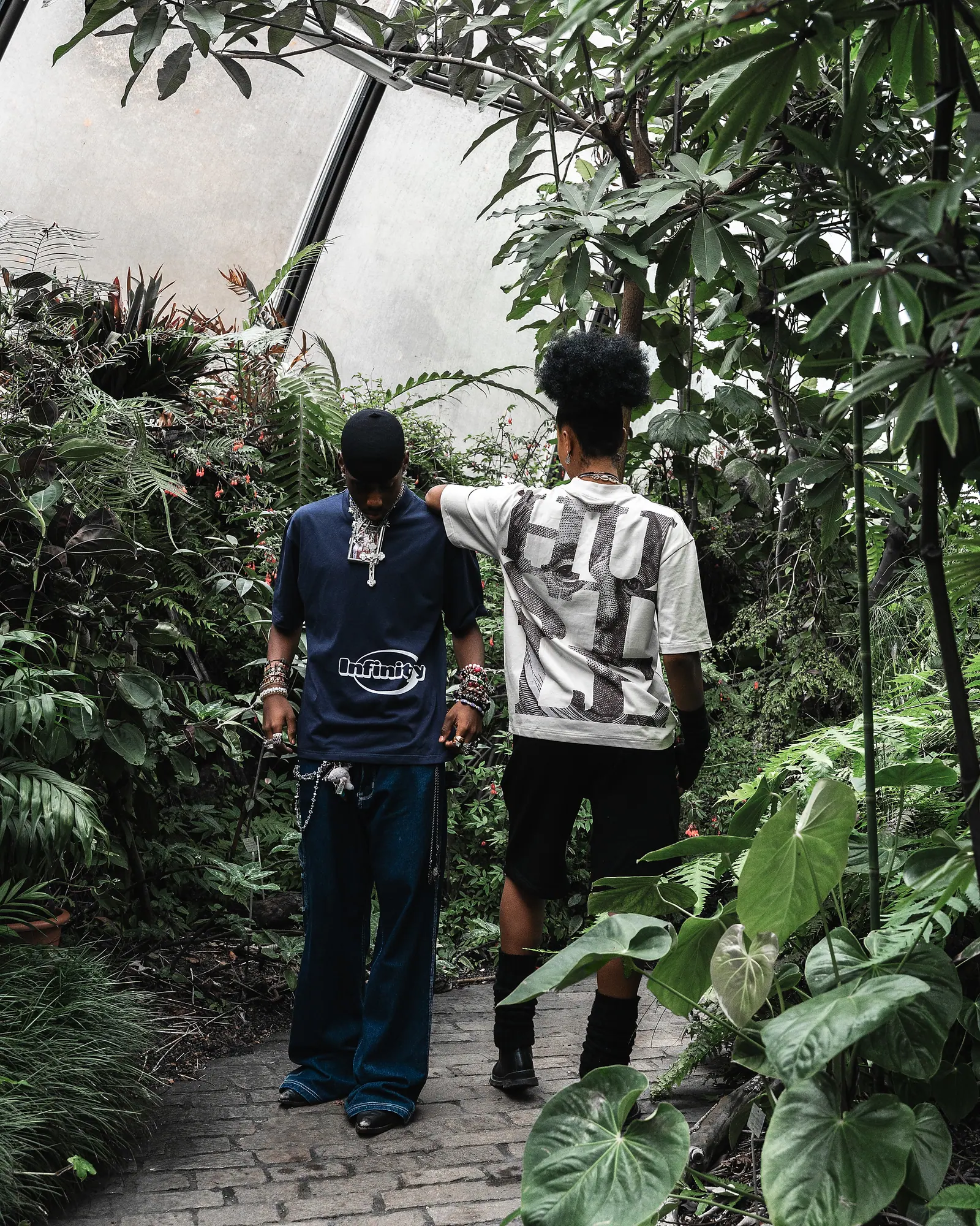 Two men on a greenhouse path, one in a navy Infinityy T-shirt and one in a grey T-shirt with a HUSTLE back print, surrounded by plants.
